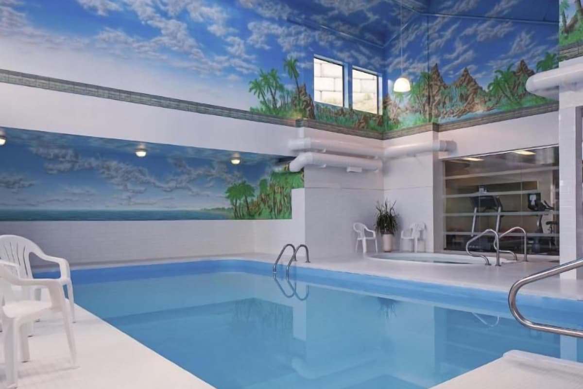 An indoor swimming pool with a ceiling mural, clear poolside chairs, and a visible exercise machine.