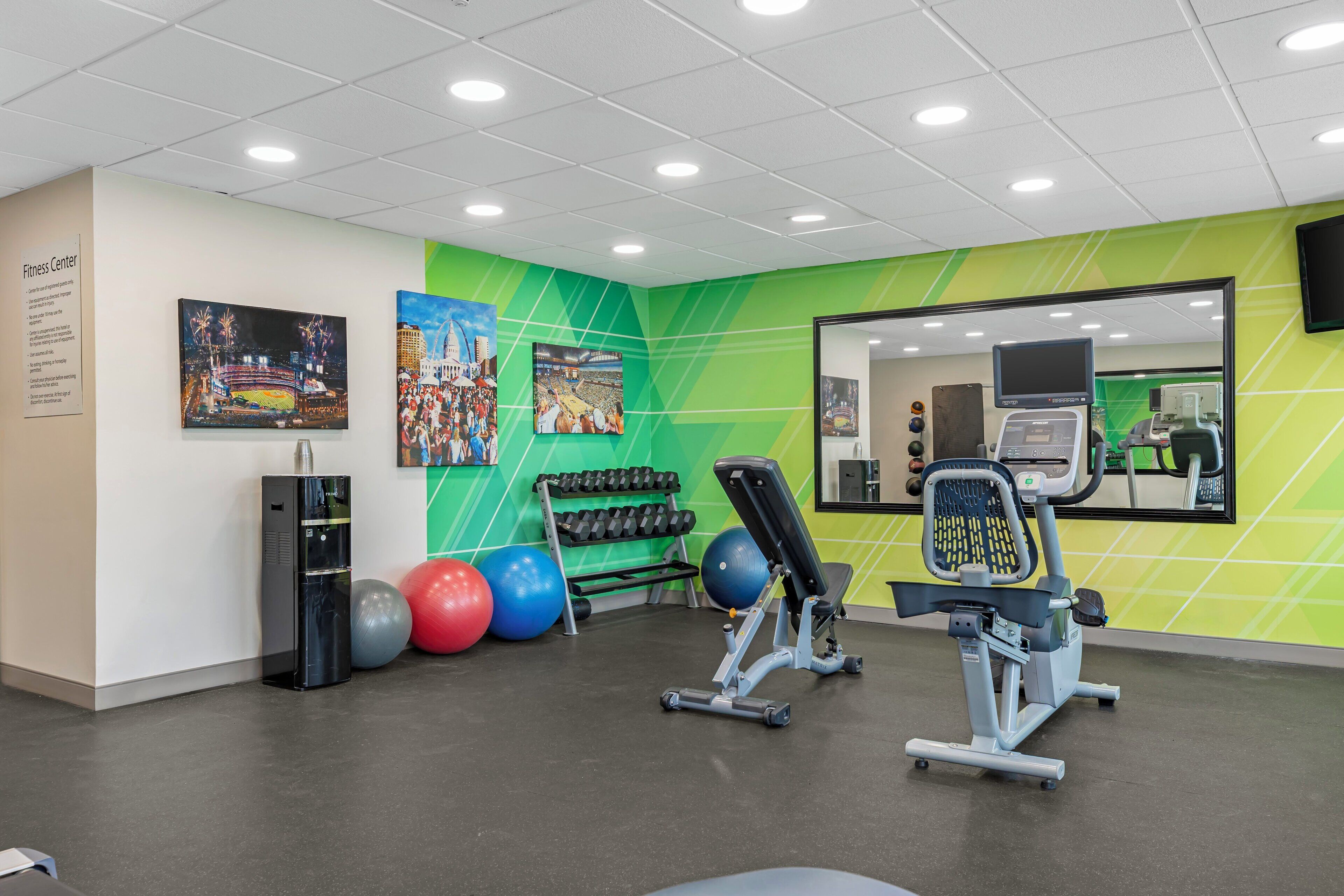 Un gimnasio con equipamiento de ejercicio, dispensador de agua y una pared decorada con motivos deportivos.