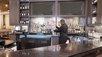 Un espace bar avec un barman préparant des boissons, des étagères remplies de bouteilles et un écran d’ordinateur posé sur le comptoir.