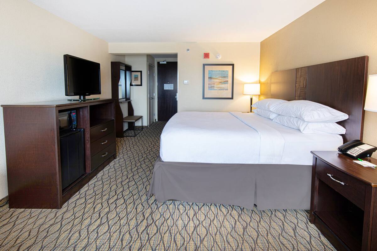 A hotel room with a bed, a desk with a television, a bedside table with a lamp, and a door leading to a bathroom.