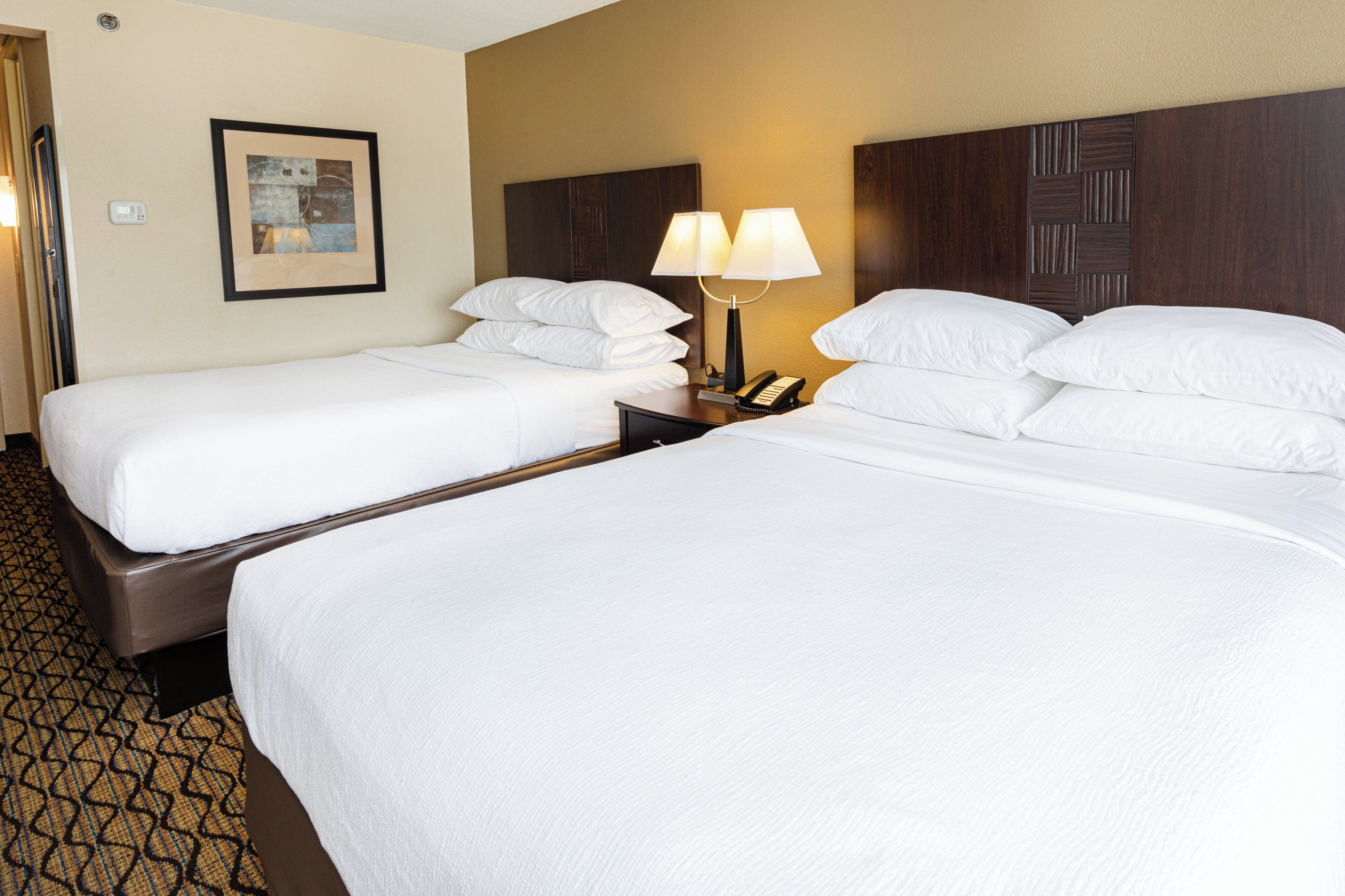 A hotel room with two beds, a nightstand with a lamp, a framed picture on the wall, and a patterned carpet.