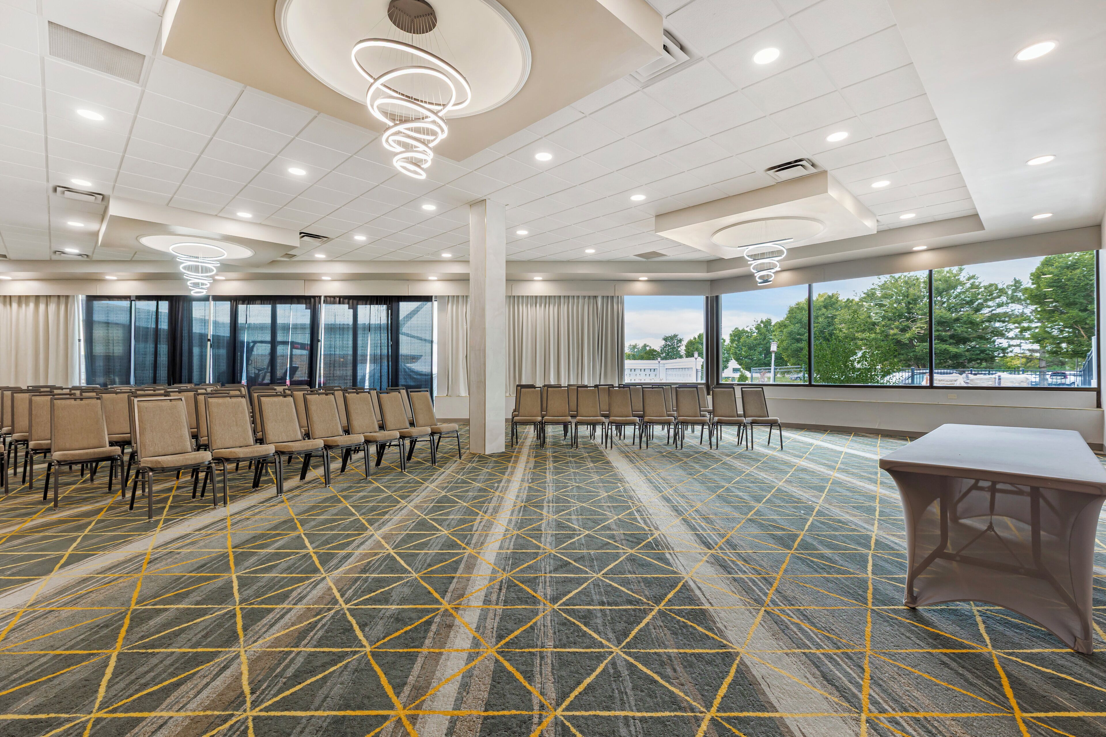 A conference room with rows of chairs, a patterned carpet, and large windows offering a view of the outdoors.