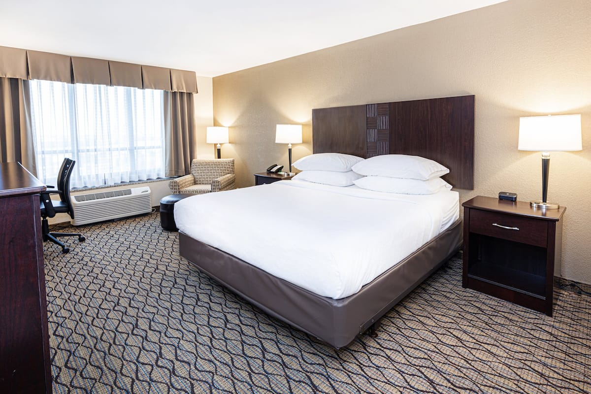 A hotel room with a large bed, two bedside tables with lamps, a desk with a chair, a chair, and a large window with curtains.