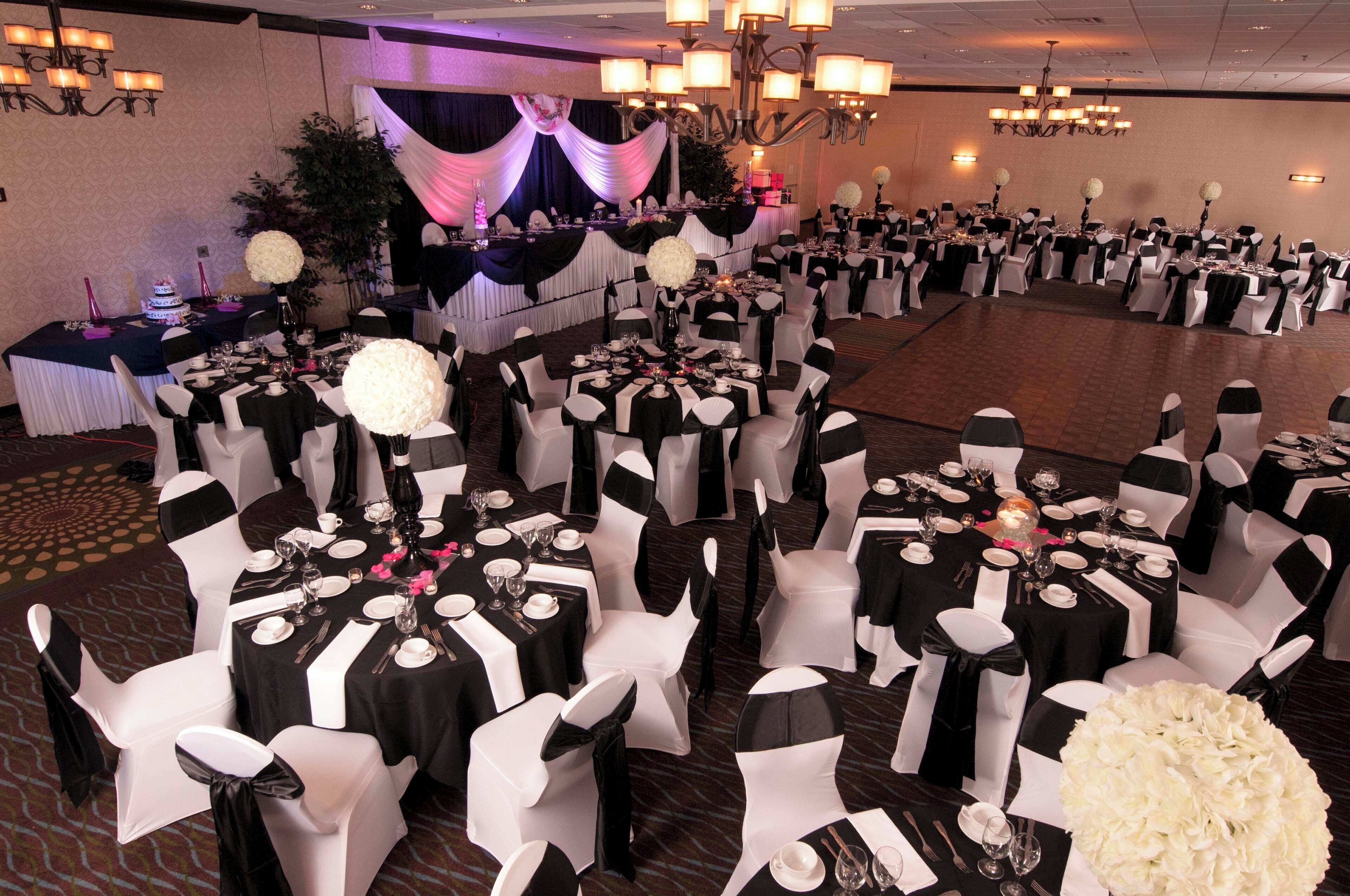 A banquet hall with round tables set for a formal event, featuring white chairs with black sashes, black tablecloths, and floral centerpieces.
