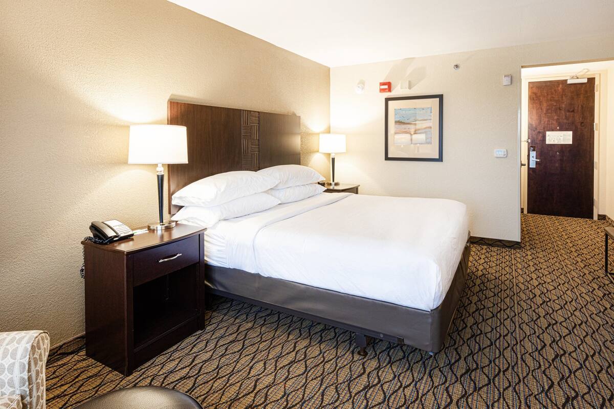A hotel room with a bed, two bedside lamps, a nightstand with a phone, a door, and a carpeted floor.