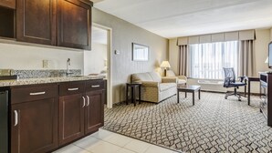 Habitación de hotel con kitchenette, sofá, escritorio y un amplio ventanal con cortinas.