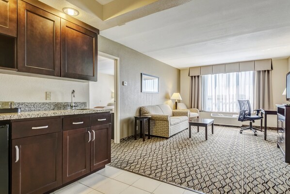 A hotel room with a kitchenette, a sofa, a desk, and a large window with curtains.