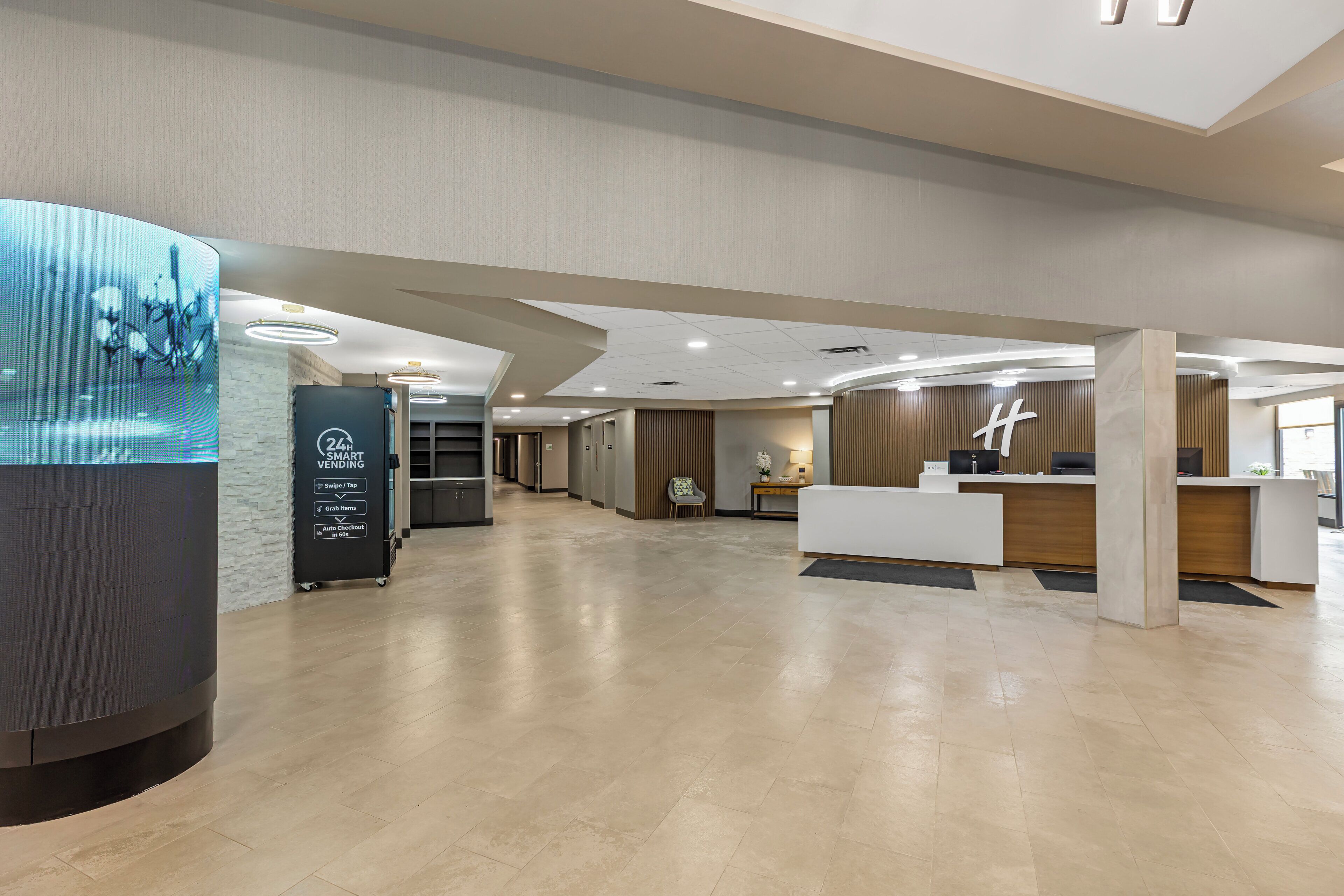 Un lobby moderno de hotel con una mesa de recepción, un quiosco de información digital y un largo pasillo que conduce a otras áreas.