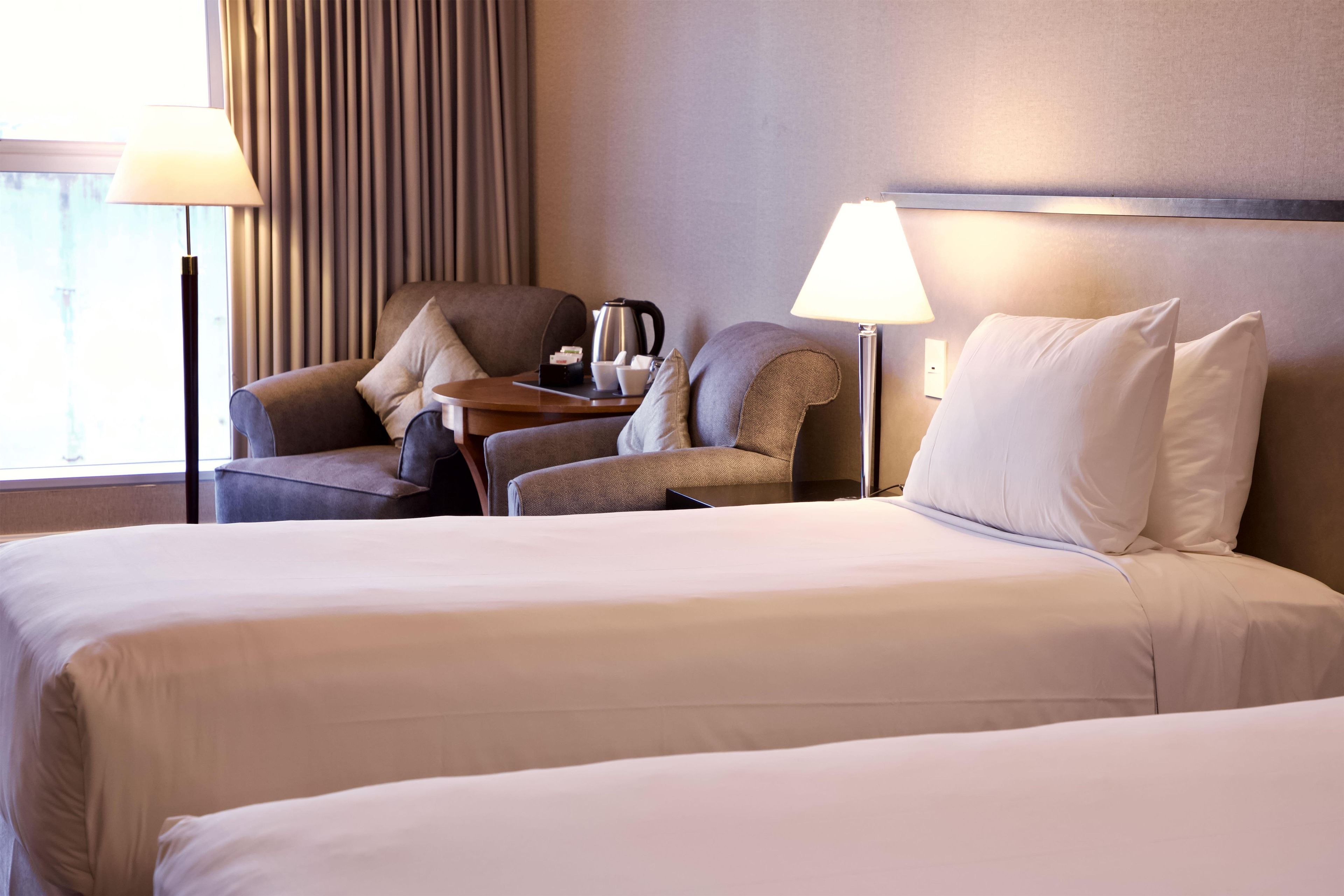 Habitación de hotel con dos camas, un área de descanso con una silla y una mesa, y una lámpara.