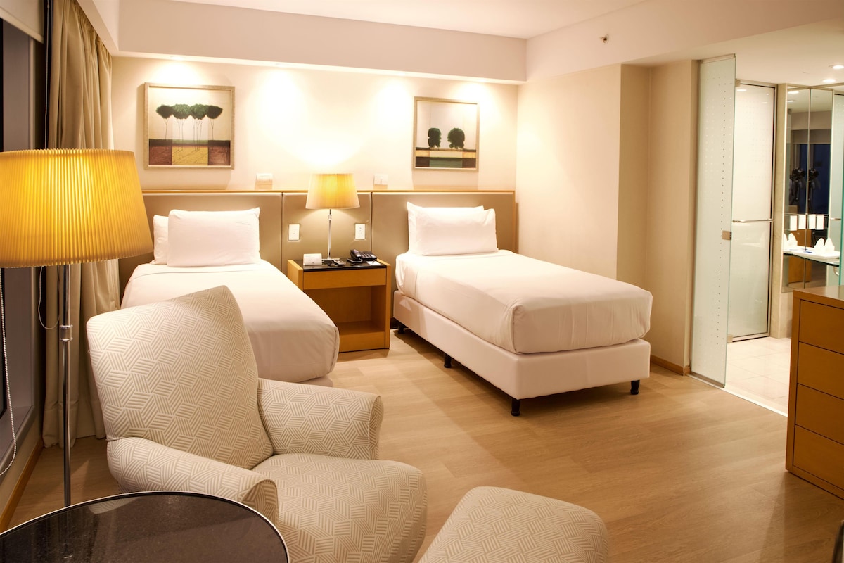 A hotel room with two beds, a desk, a chair, and a lamp.