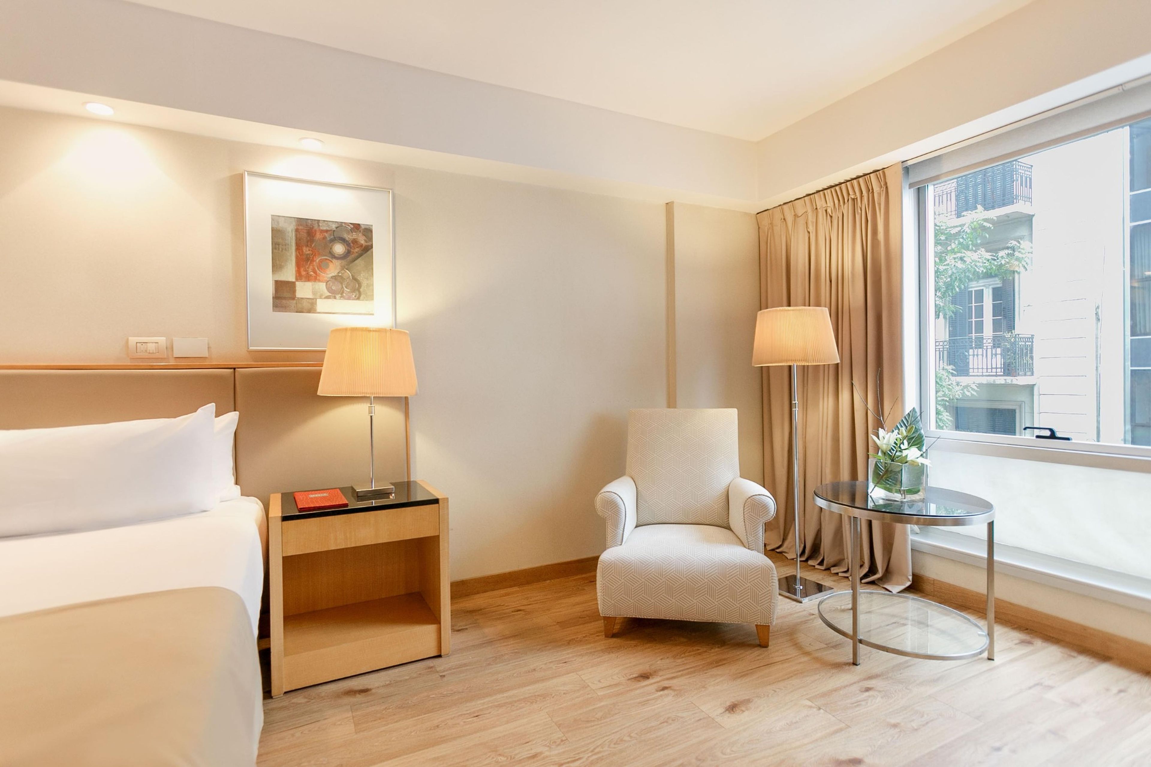 A hotel room with a bed, a chair, a nightstand, a lamp, a table with a vase, and a window with curtains.