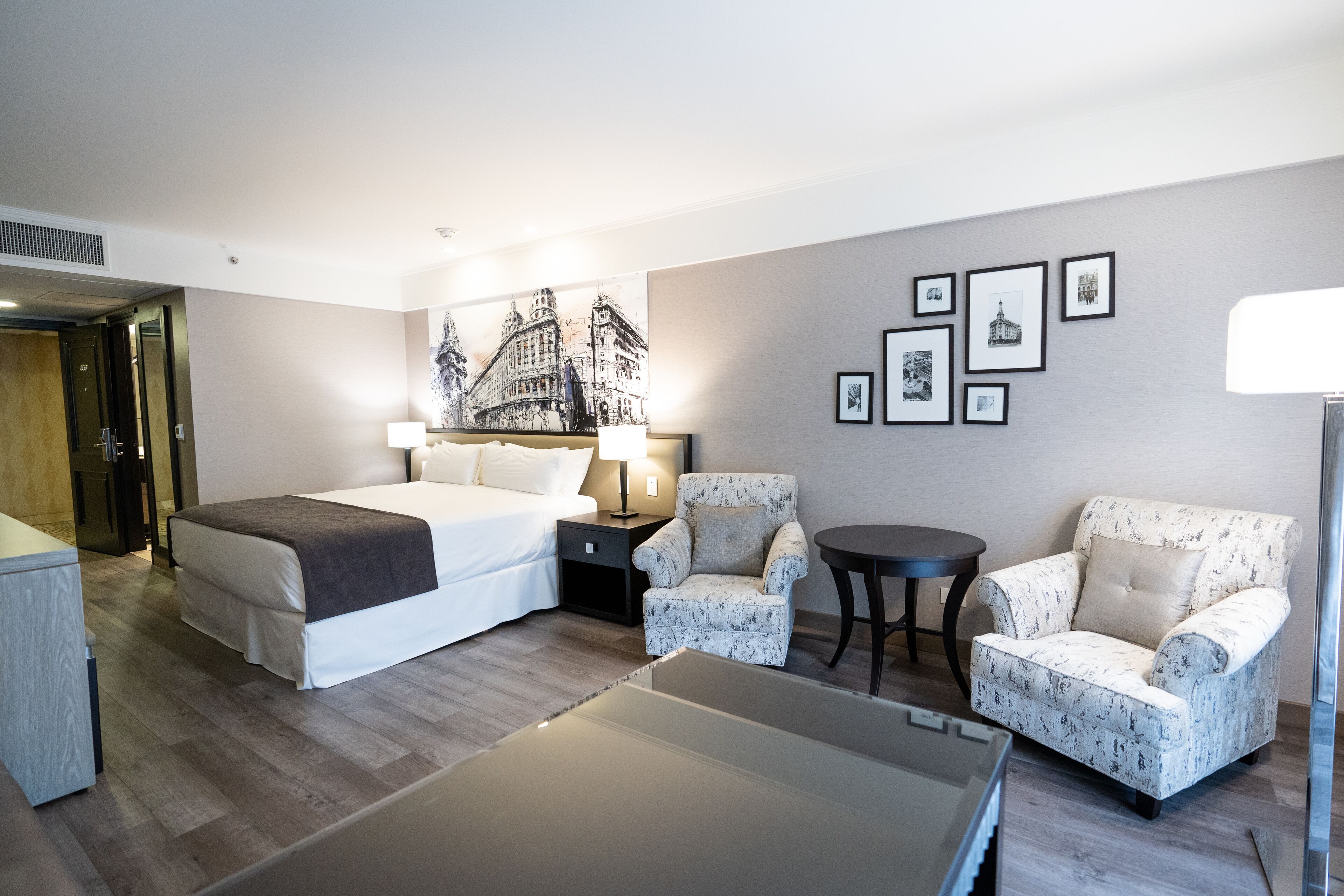 A hotel room with a bed, two armchairs, a small table, and a wall with framed pictures.