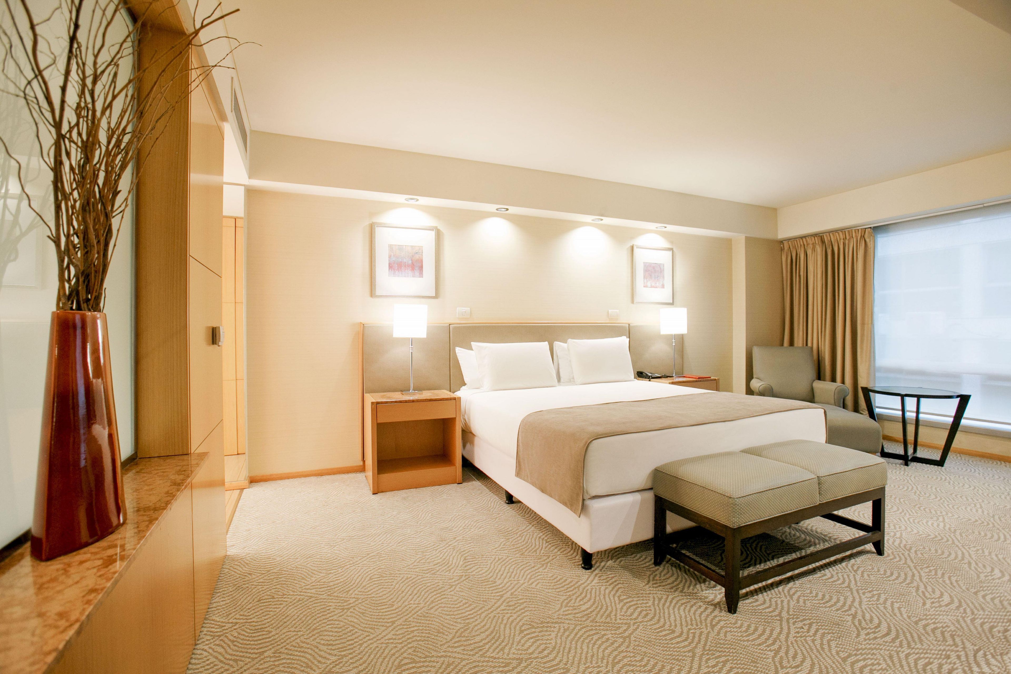 A hotel room with a bed, bedside tables, a chair, a small table, a vase with branches, and a window with curtains.