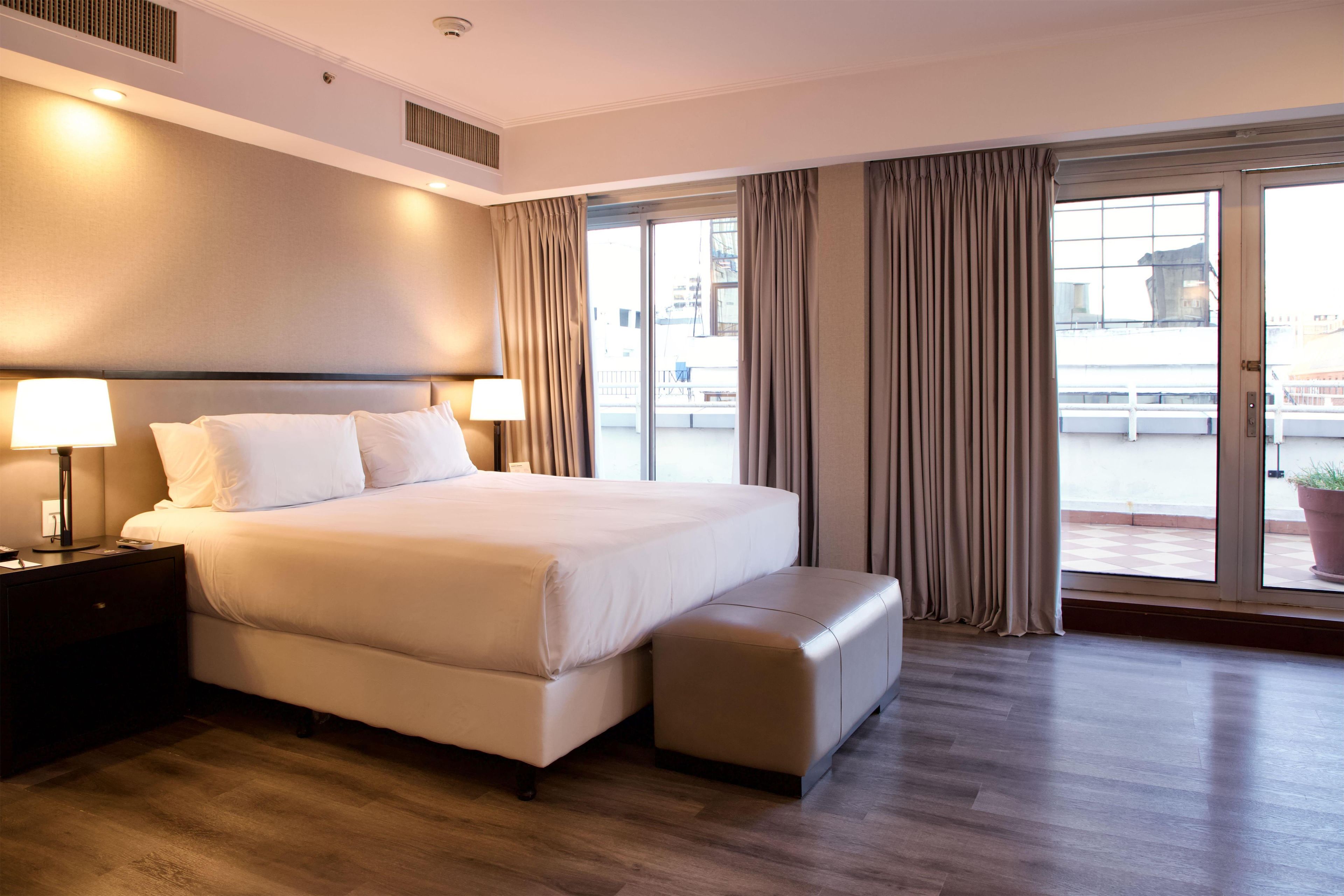 A hotel room with a large bed, bedside lamps, a small table, and a view of the city through the window.