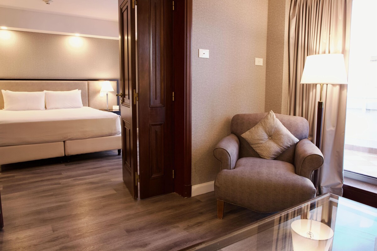 Een hotelkamer met een bed, een stoel, een glazen tafel en een raam met gordijnen.