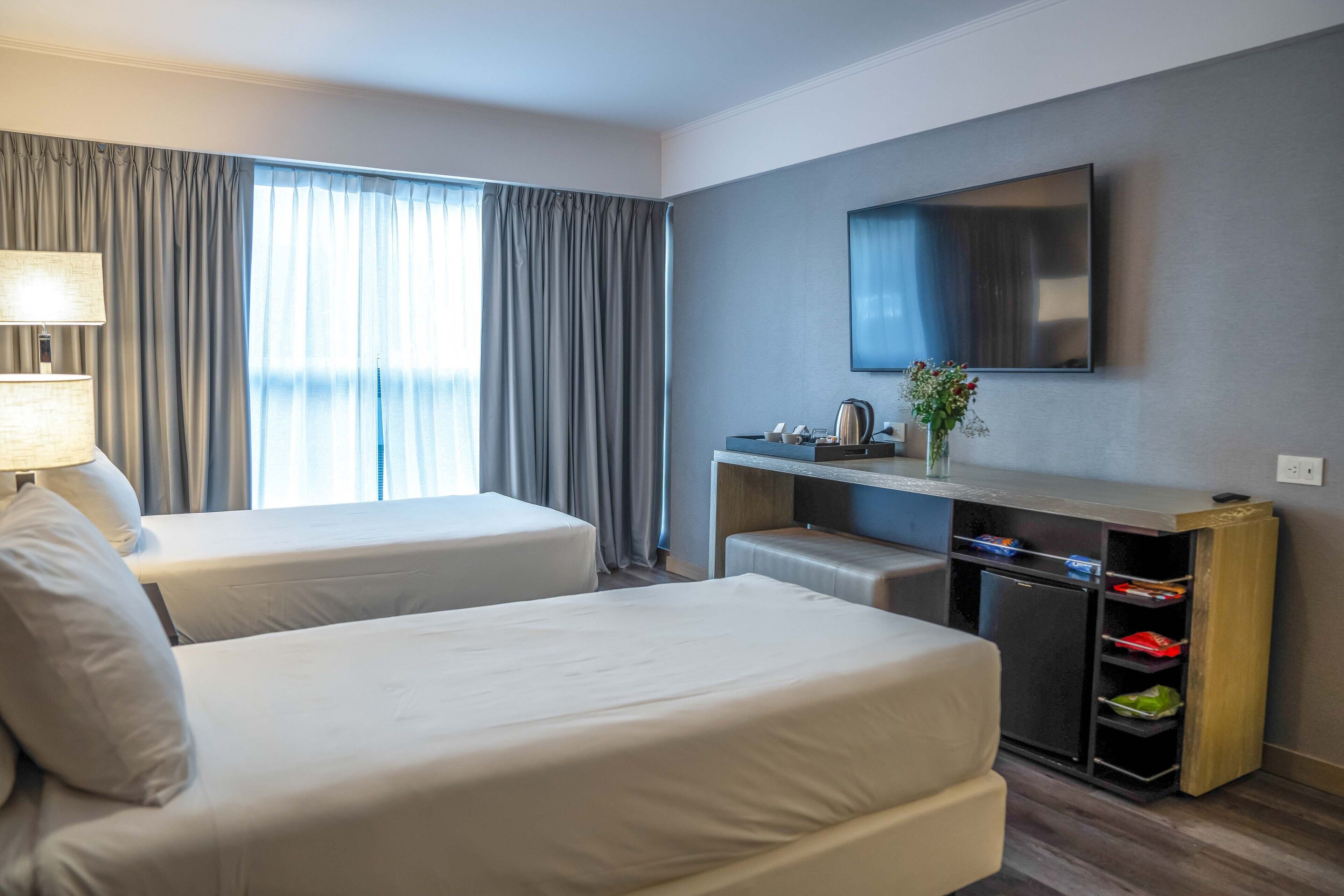 A hotel room with two beds, a desk with a flat-screen TV, a lamp, and a small cabinet with storage.