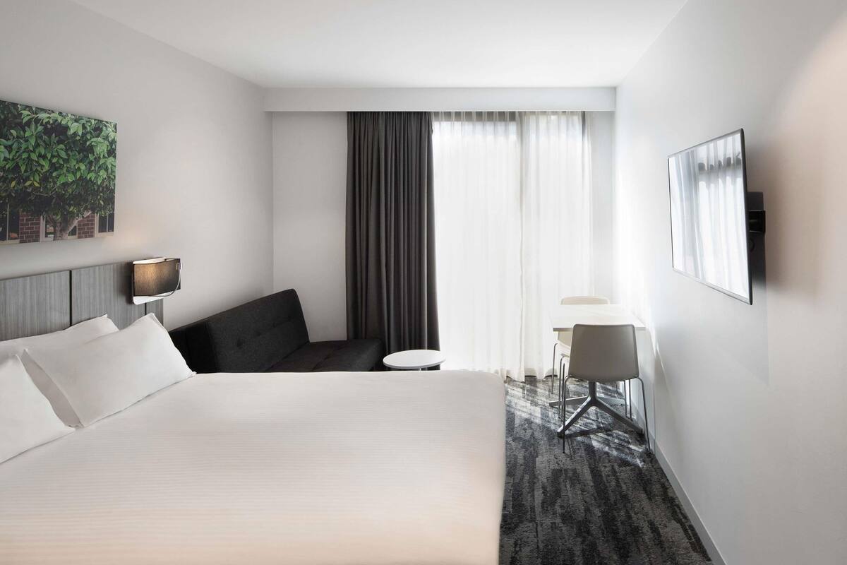 Ein Hotelzimmer mit einem Bett, einer Sitzecke mit Stuhl und kleinem Tisch, einem an der Wand befestigten Fernseher und einem großen Fenster mit Vorhängen.