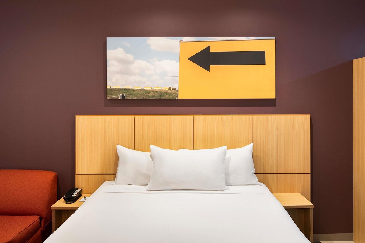 Una habitación de hotel con una cama, una mesita de noche, una silla roja, un cabecero de madera y un letrero en la pared con una flecha.