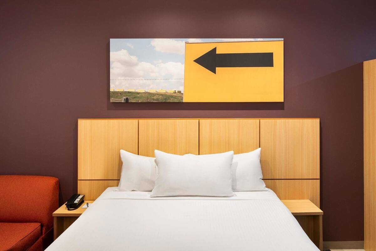 Ein Hotelzimmer mit einem Bett, einem Nachttisch, einem roten Sessel, einem Holz-Kopfteil und einem an der Wand befestigten Schild mit einem Pfeil.