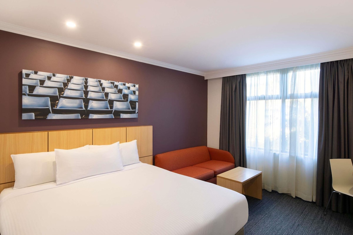 Habitación de hotel con una cama grande, un sofá, una mesita y una obra de arte en la pared, además de un ventanal con cortinas.