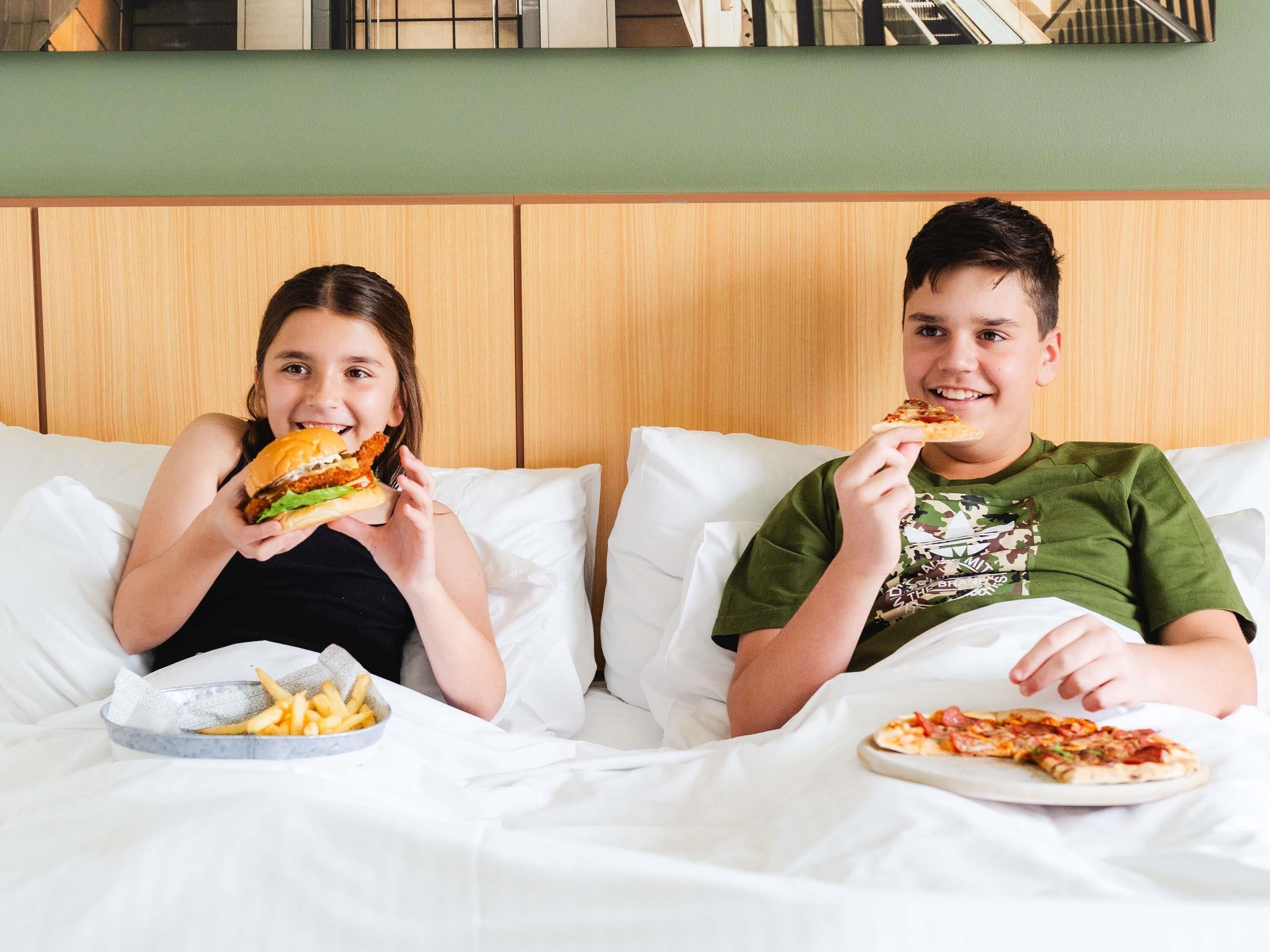 Twee personen zitten op een bed met een bord pizza en een schaal friet.