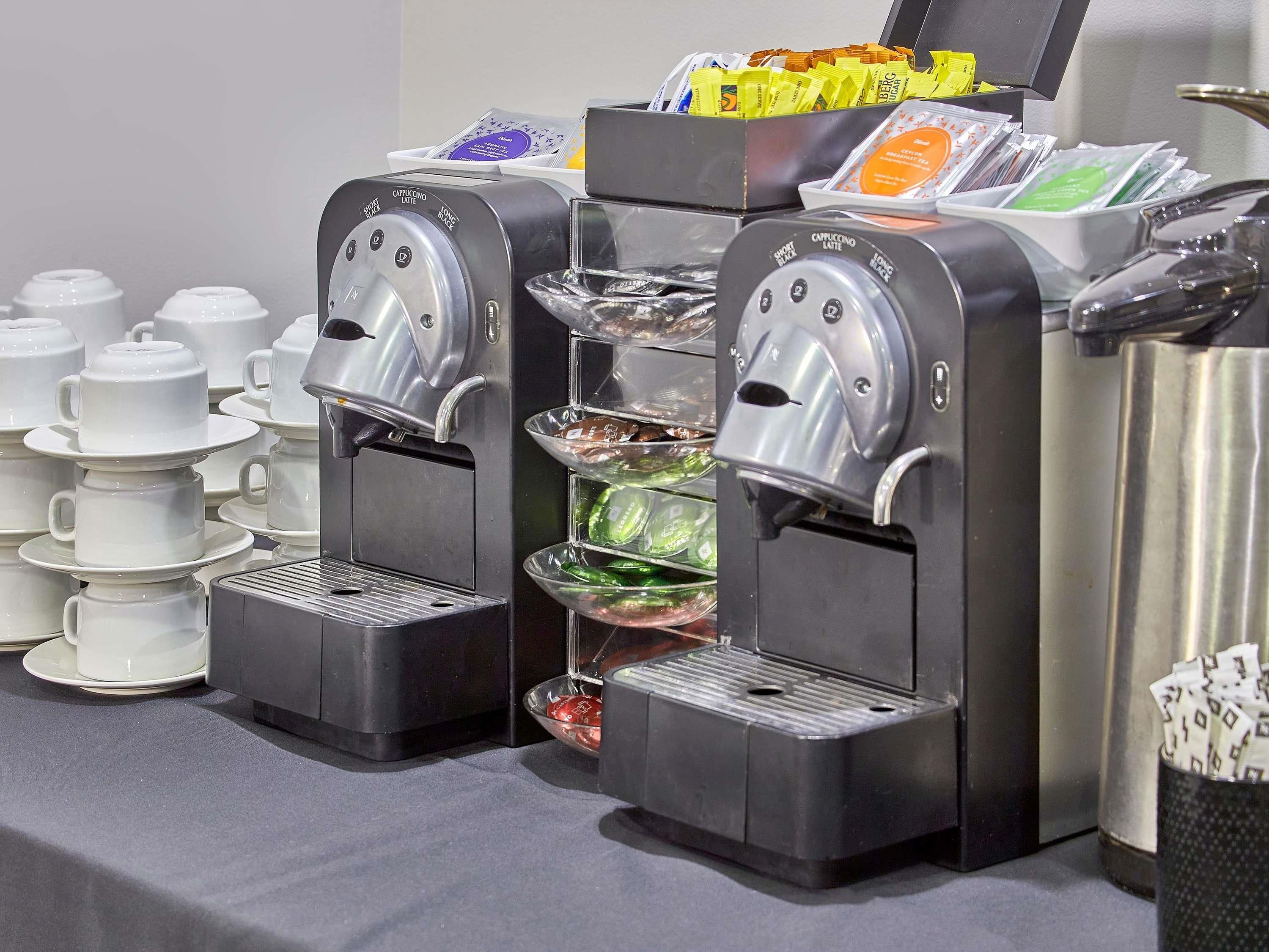 A coffee station with multiple coffee dispensers, a variety of coffee pods, and a stack of white cups.