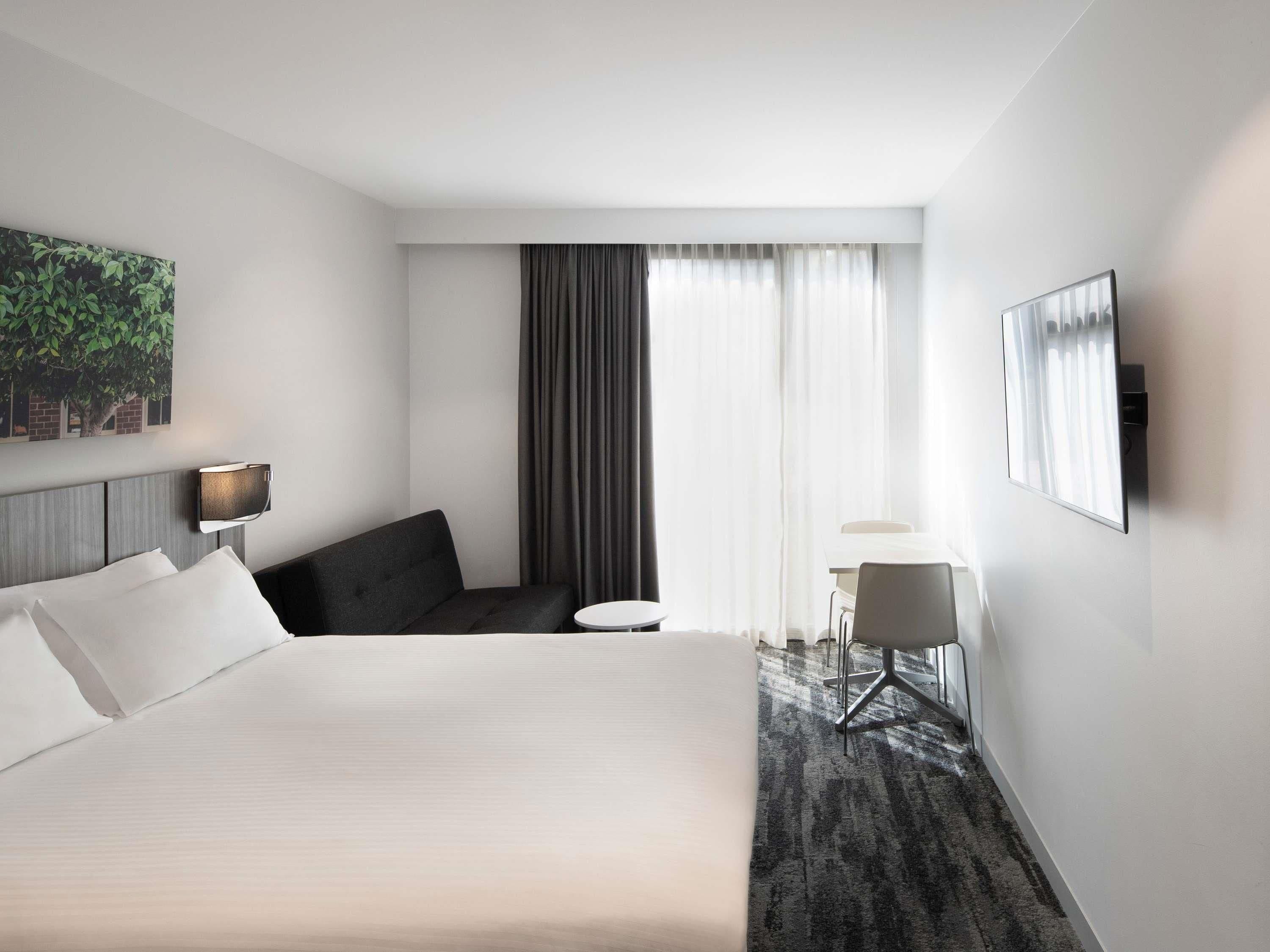 Una camera d'albergo con un letto, un'area salotto con una poltrona e un tavolino, una TV a parete e un'ampia finestra con tende.