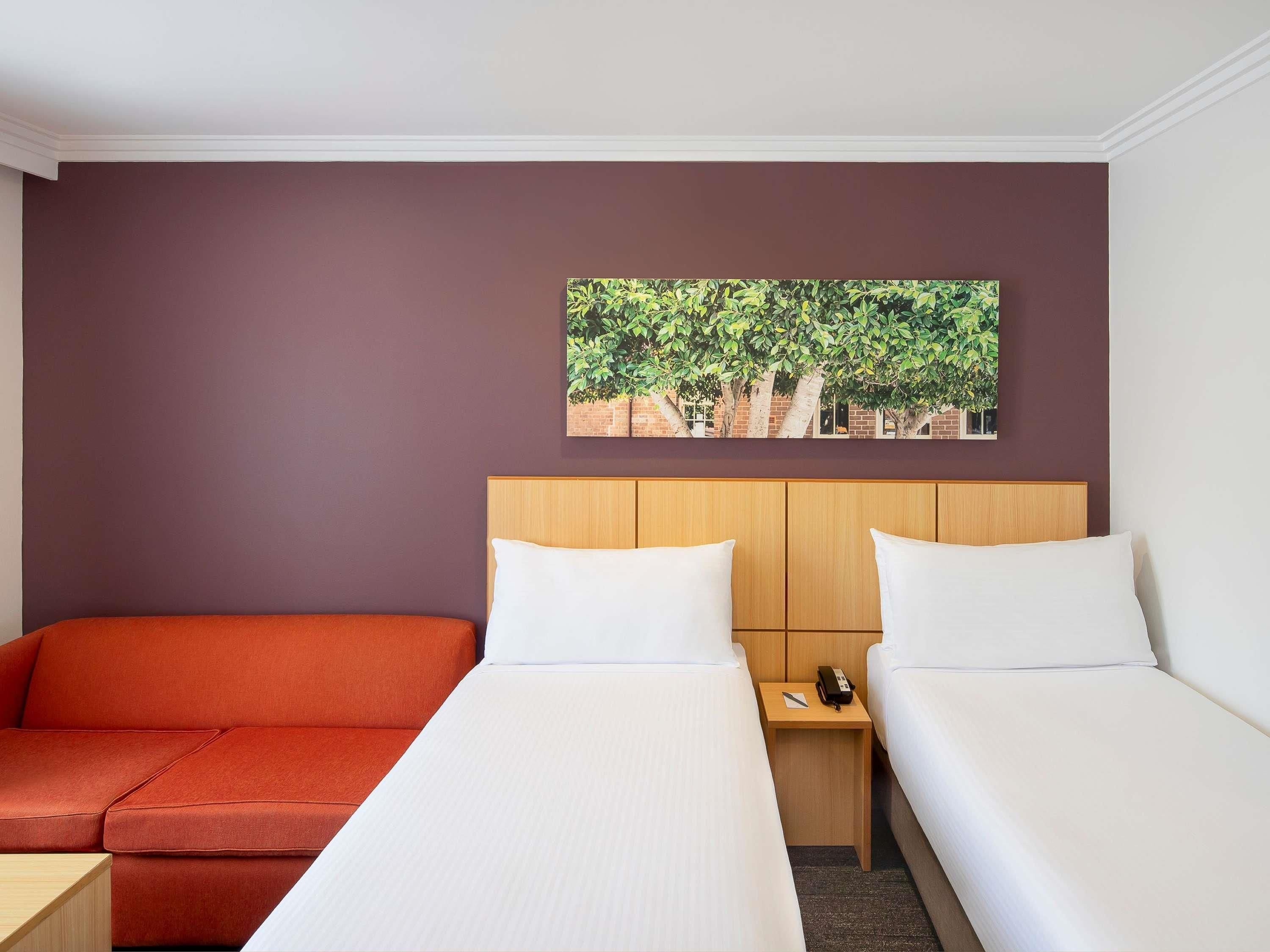 A hotel room with two beds, a sofa, and a painting of trees on the wall.