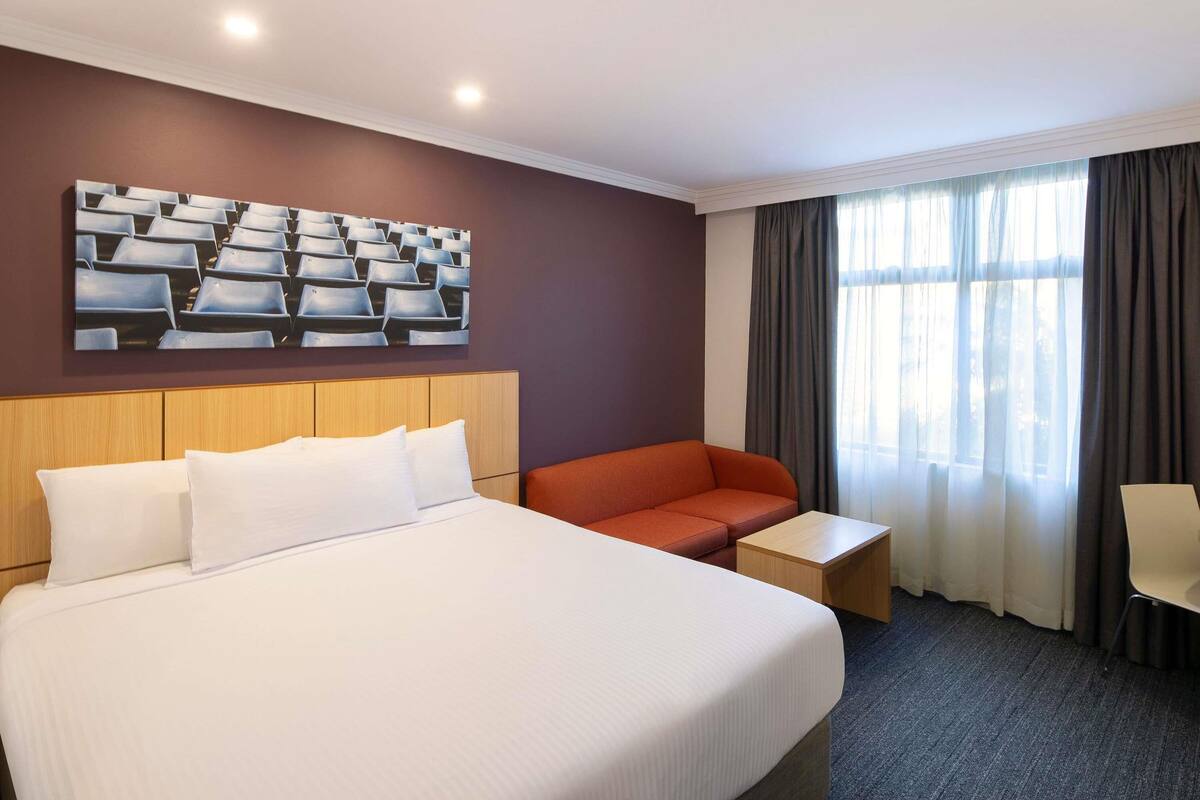 Ein Hotelzimmer mit einem großen Bett, einem Sofa, einem kleinen Tisch, einem Wandbild und einem Fenster mit Vorhängen.