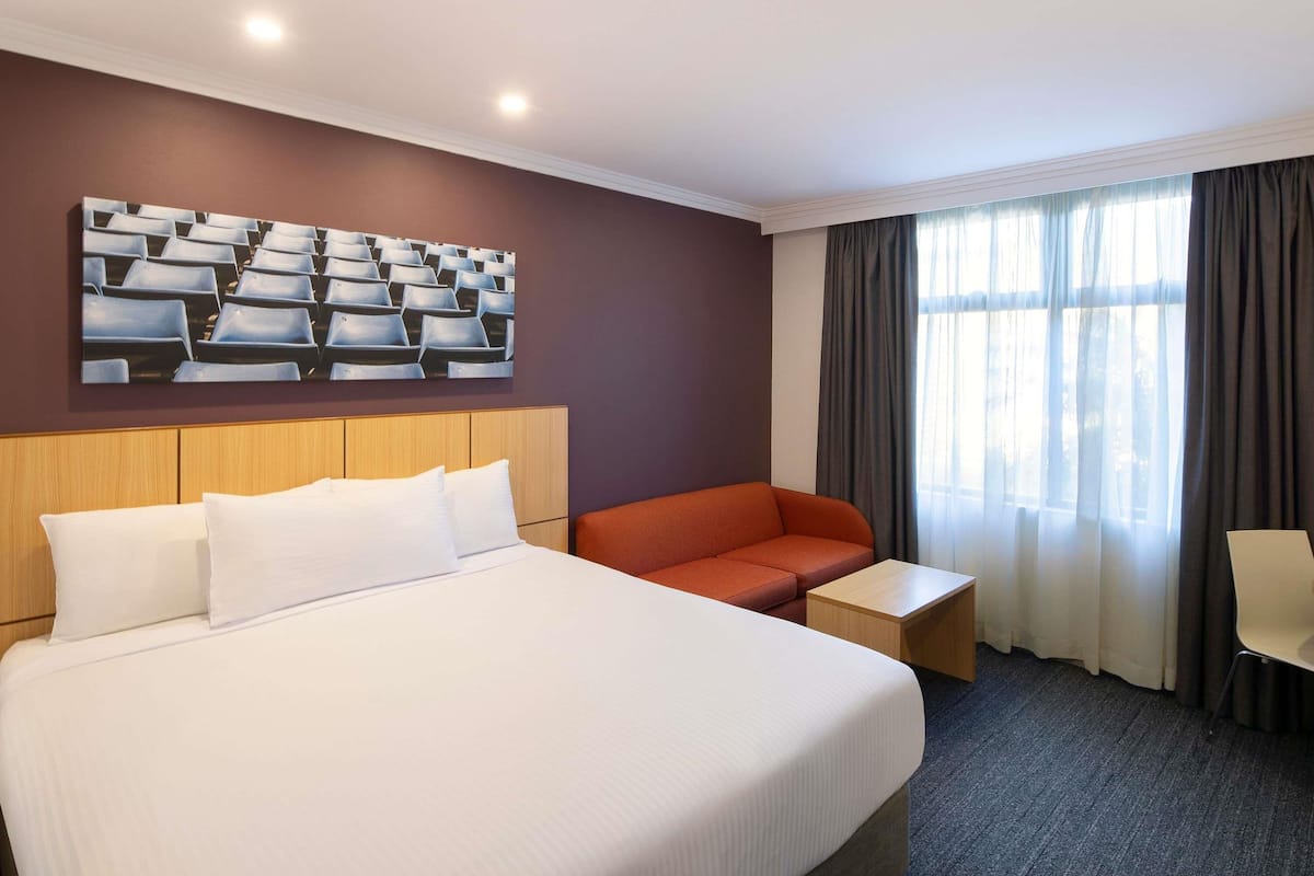 A hotel room with a large bed, a sofa, a small table, a wall-mounted artwork, and a window with curtains.