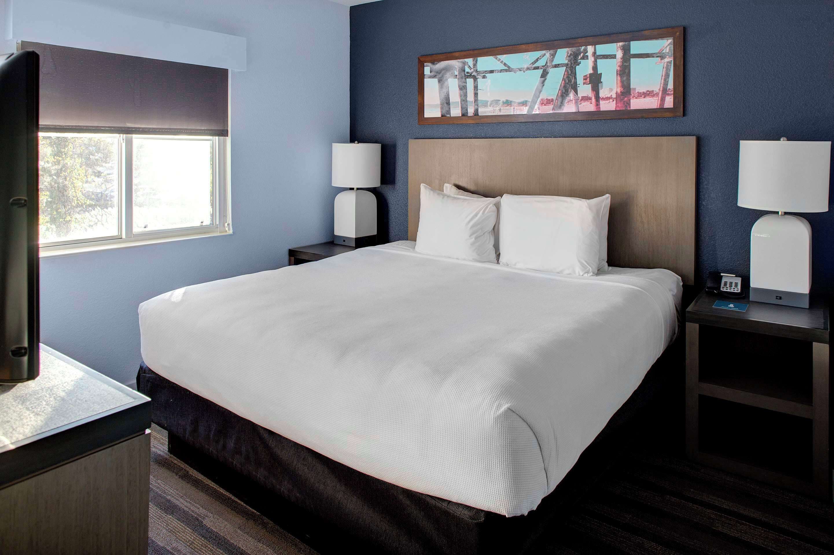 A hotel room with a large bed, two bedside tables with lamps, a painting on the wall, and a window with blinds.