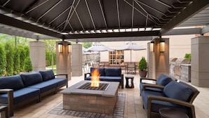 Een terras buiten met een vuurkuil, banken en parasols.