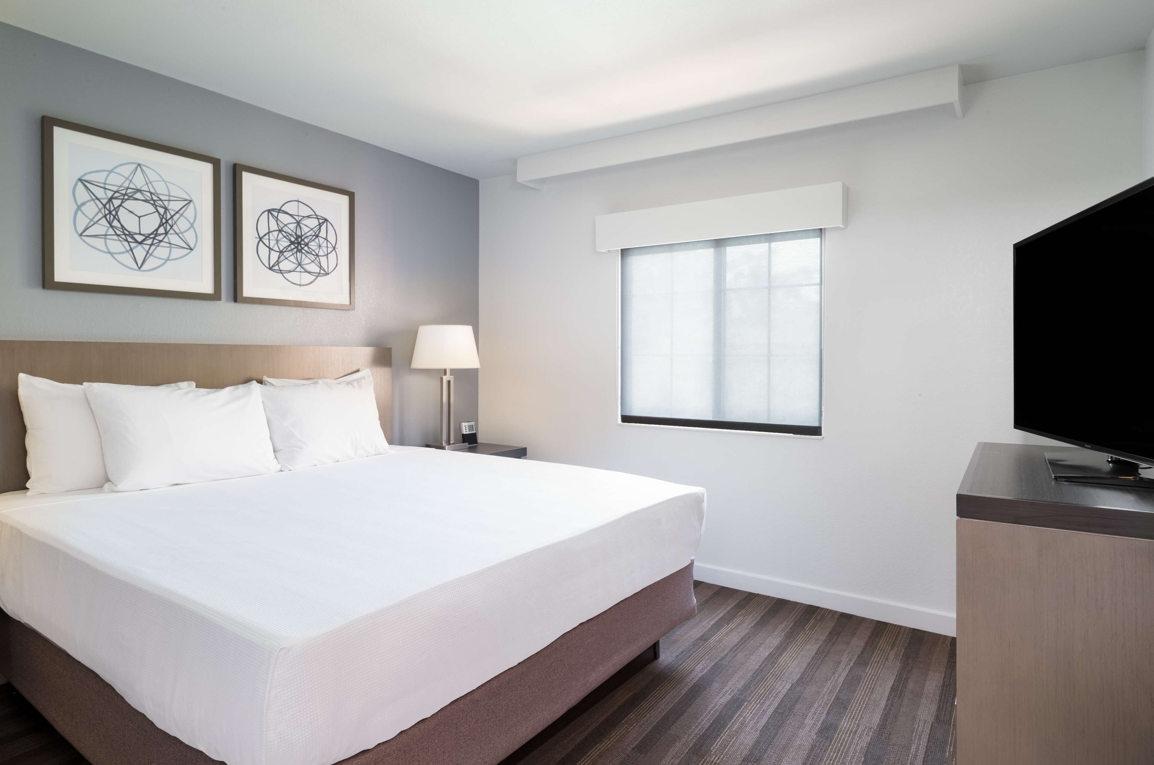 A hotel room with a large bed, a desk with a TV, a window, and two framed artworks on the wall.
