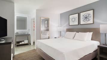 A modern hotel room with a large bed, a TV, a desk with a computer, and a bathroom with a mirror and sink.