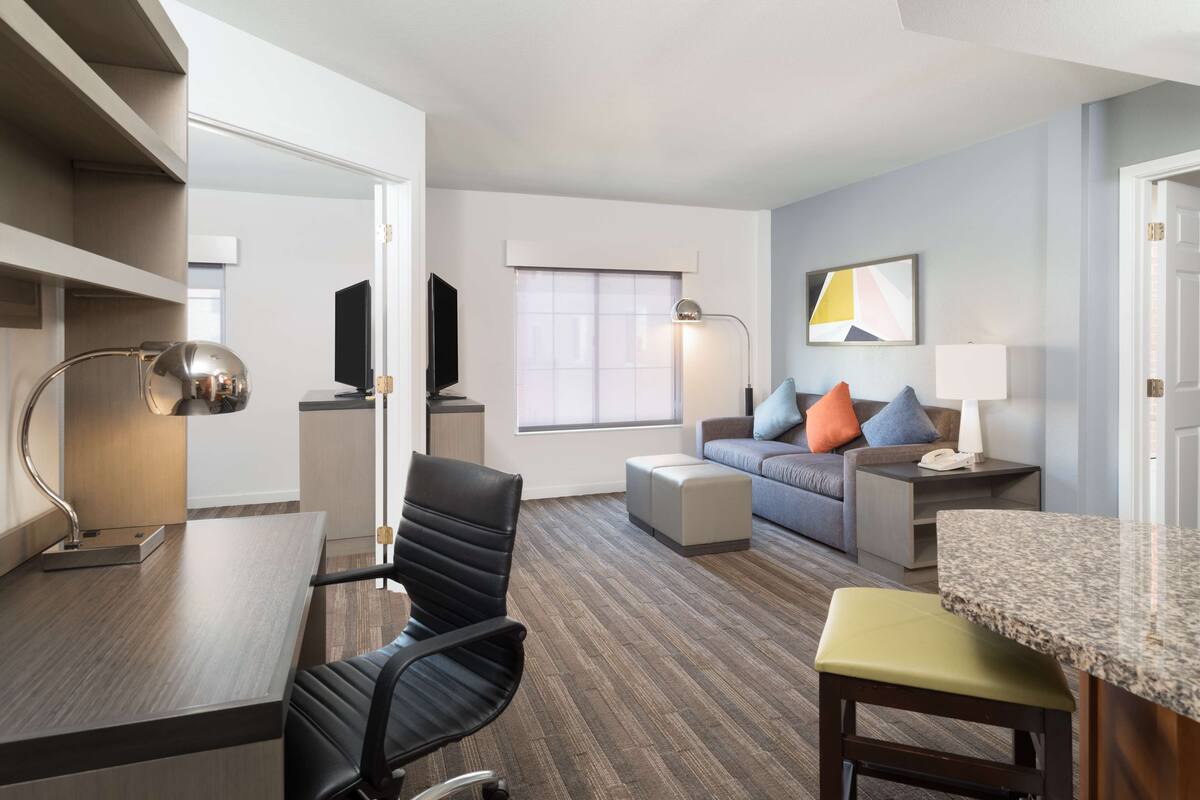 Een moderne hotelkamer met een zithoek, een bank met kleurrijke kussens, een televisie en een groot raam.