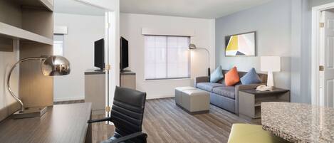 Ein modernes Hotelzimmer mit Essbereich, einer Couch mit bunten Kissen, einem Fernseher und einem großen Fenster.