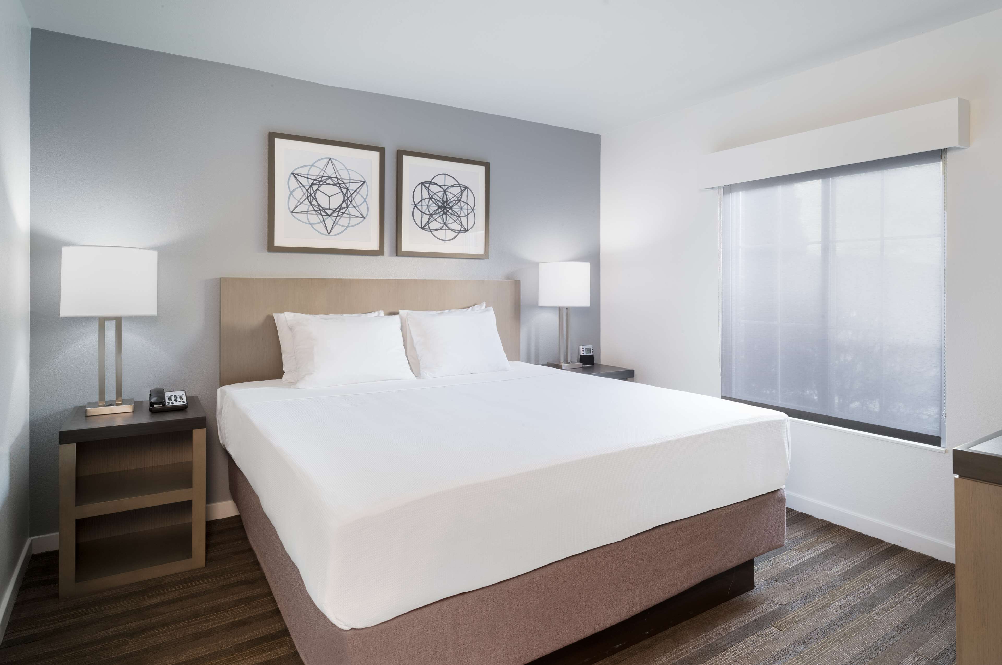 A hotel room with a large bed, two bedside tables with lamps, a window with blinds, and two framed abstract wall art pieces.