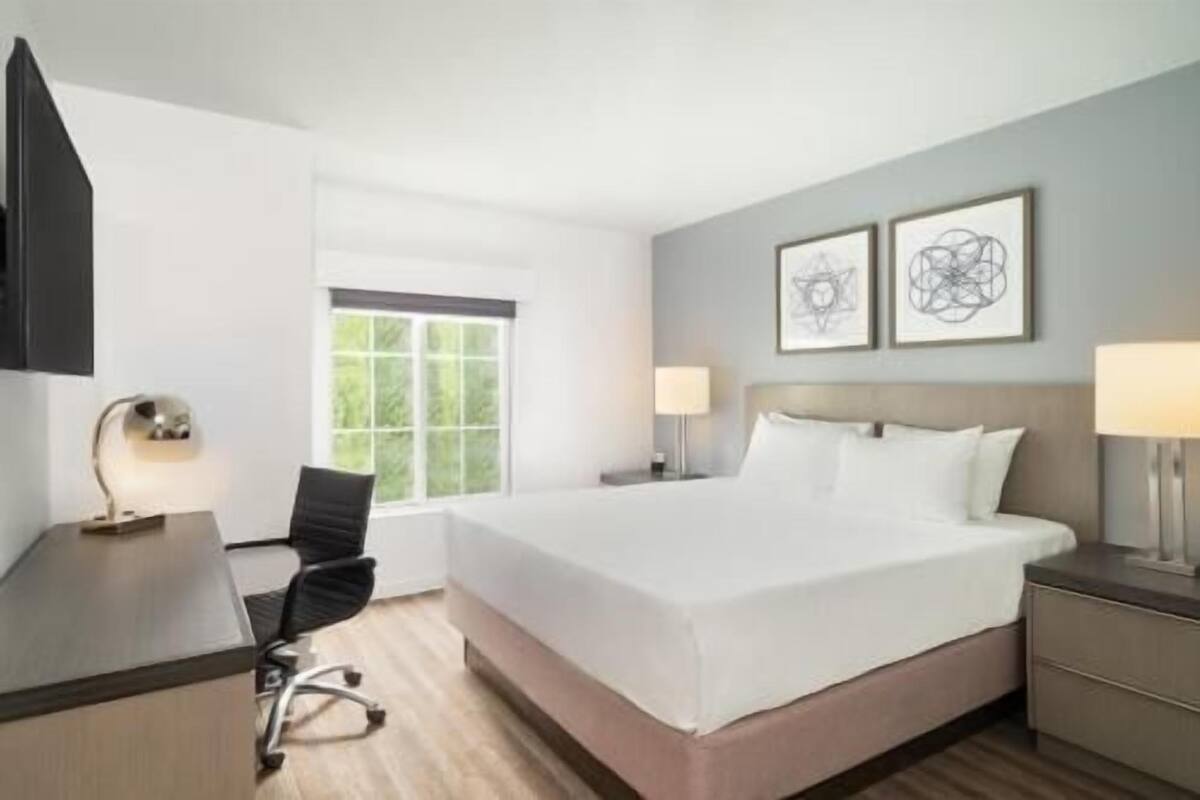 Ein Hotelzimmer mit Bett, Schreibtisch, Stuhl, Nachttischen, Lampen und Kunstwerken an den Wänden.