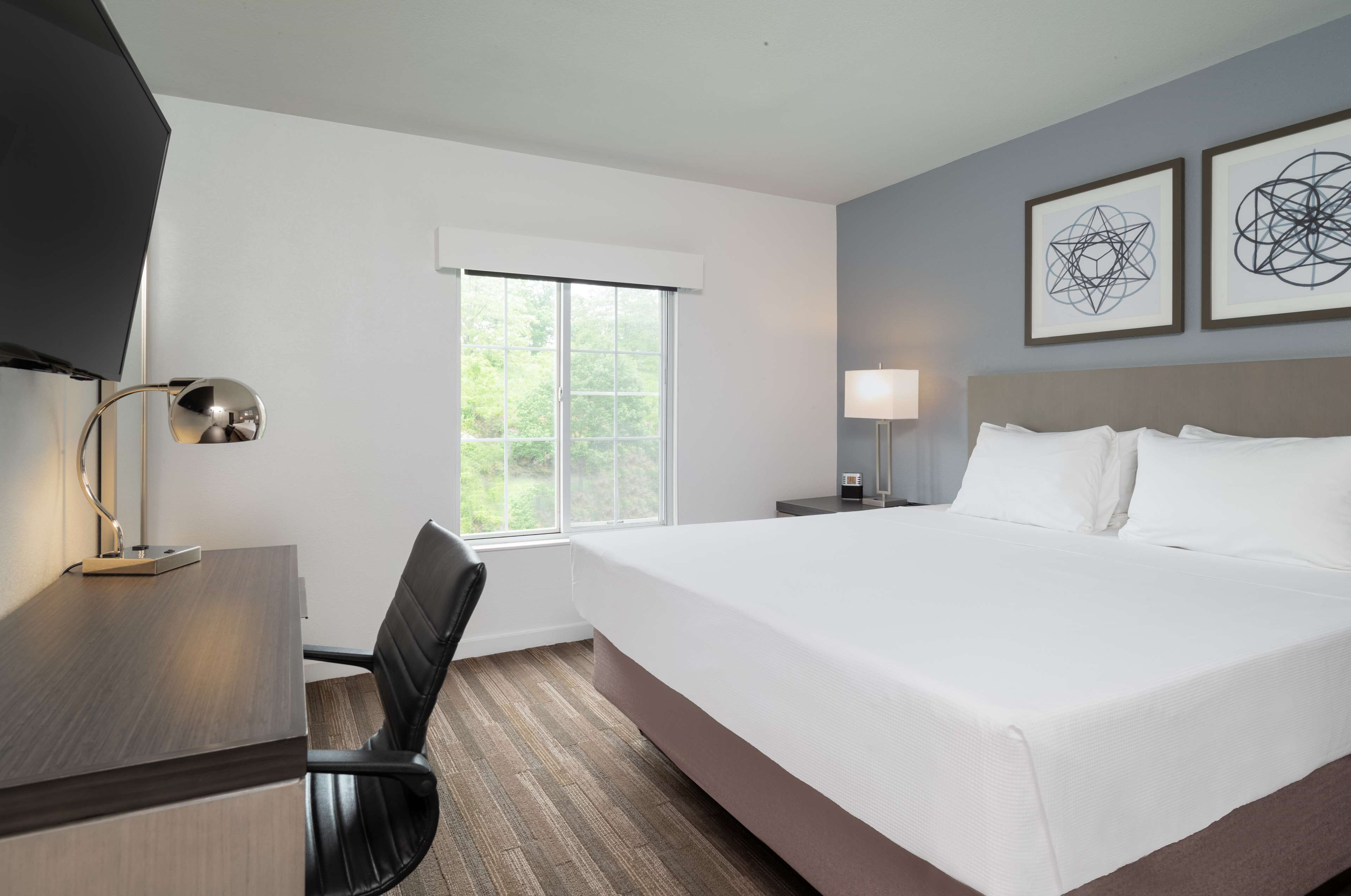 A hotel room with a large bed, a desk with a chair, a TV, and two framed artworks on the wall.