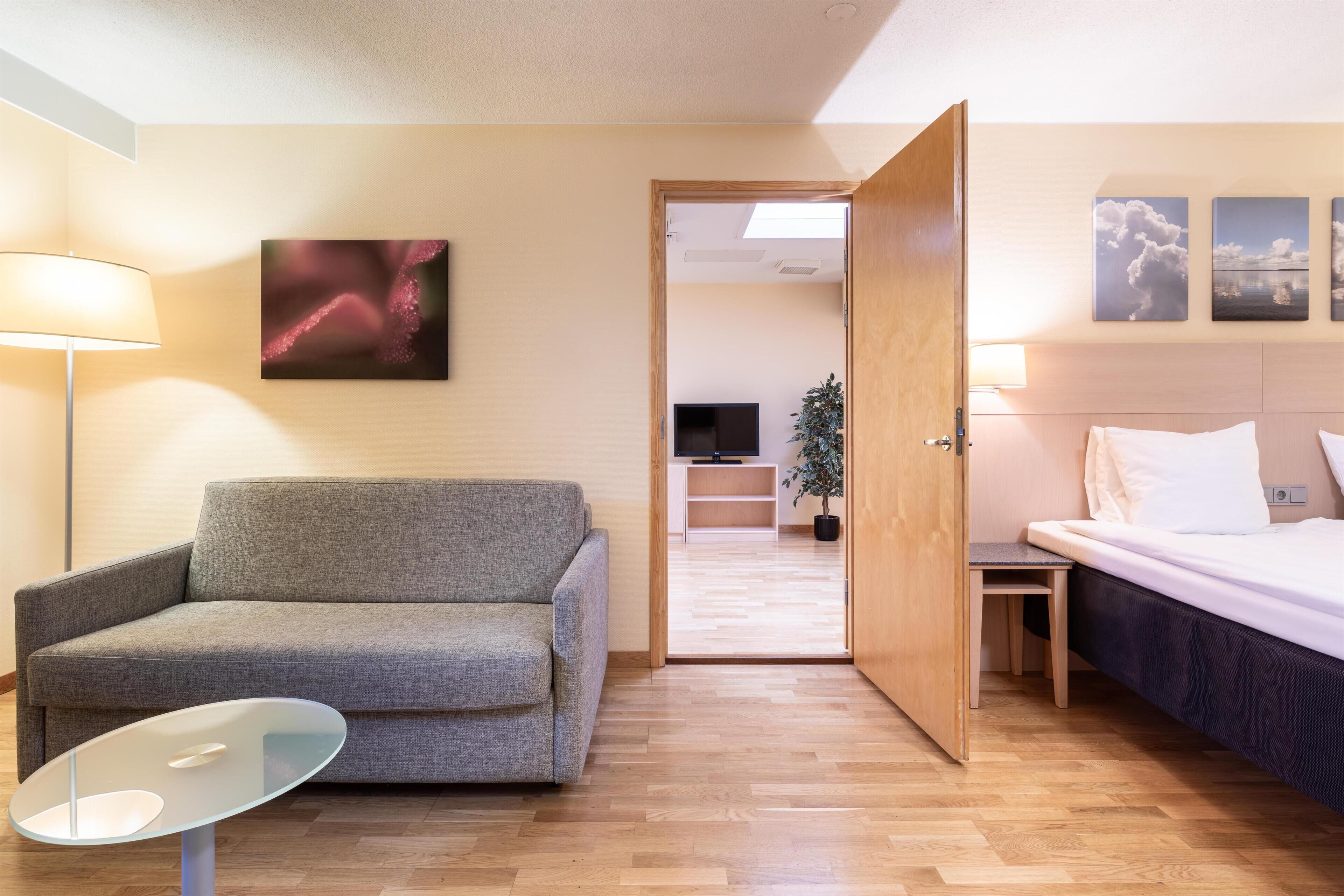 Habitación de hotel con sofá, una mesita de vidrio, cama y televisión al fondo.