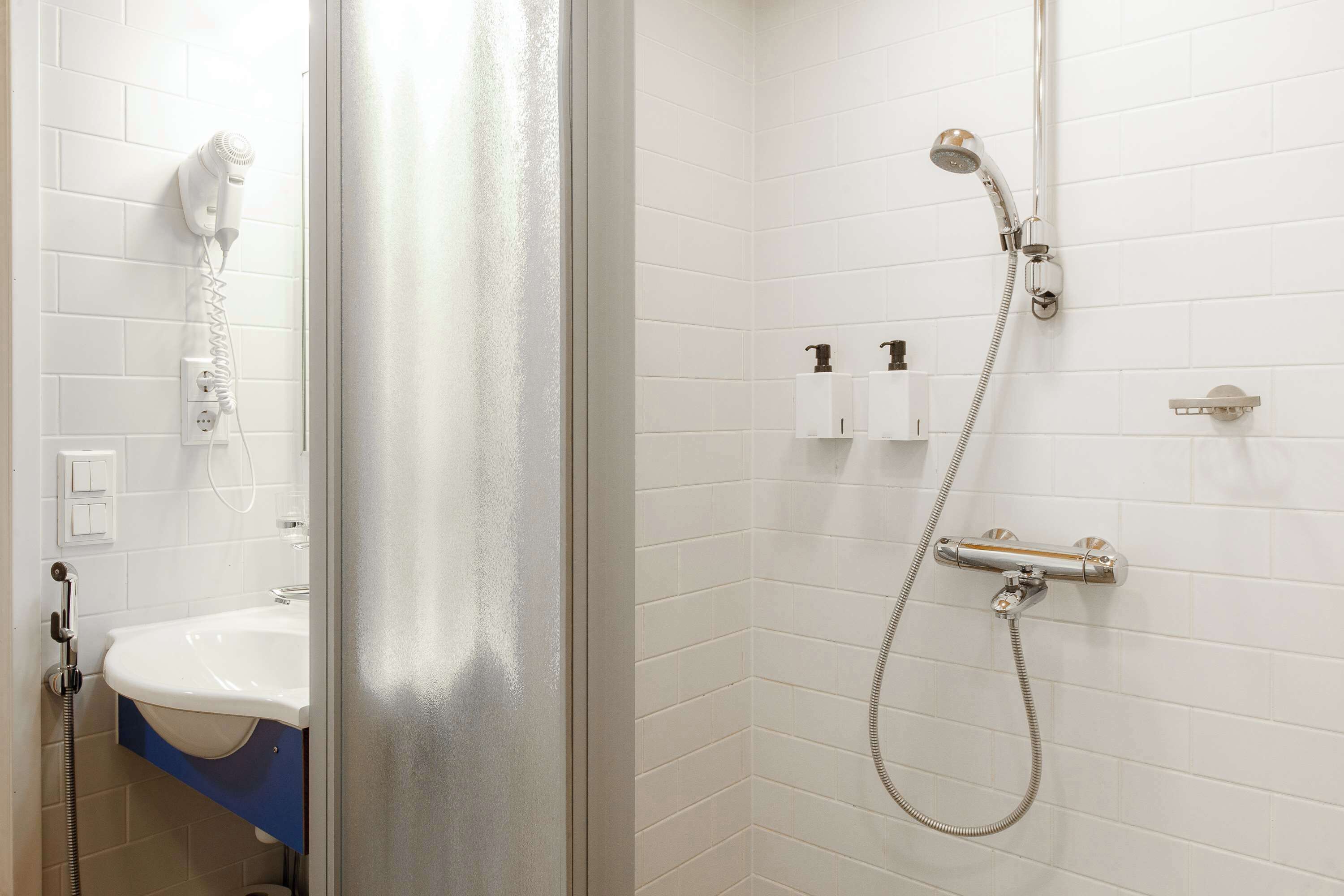 A bathroom with a white tiled wall, a sink, a mirror, a shower with a handheld showerhead, and a towel rack.