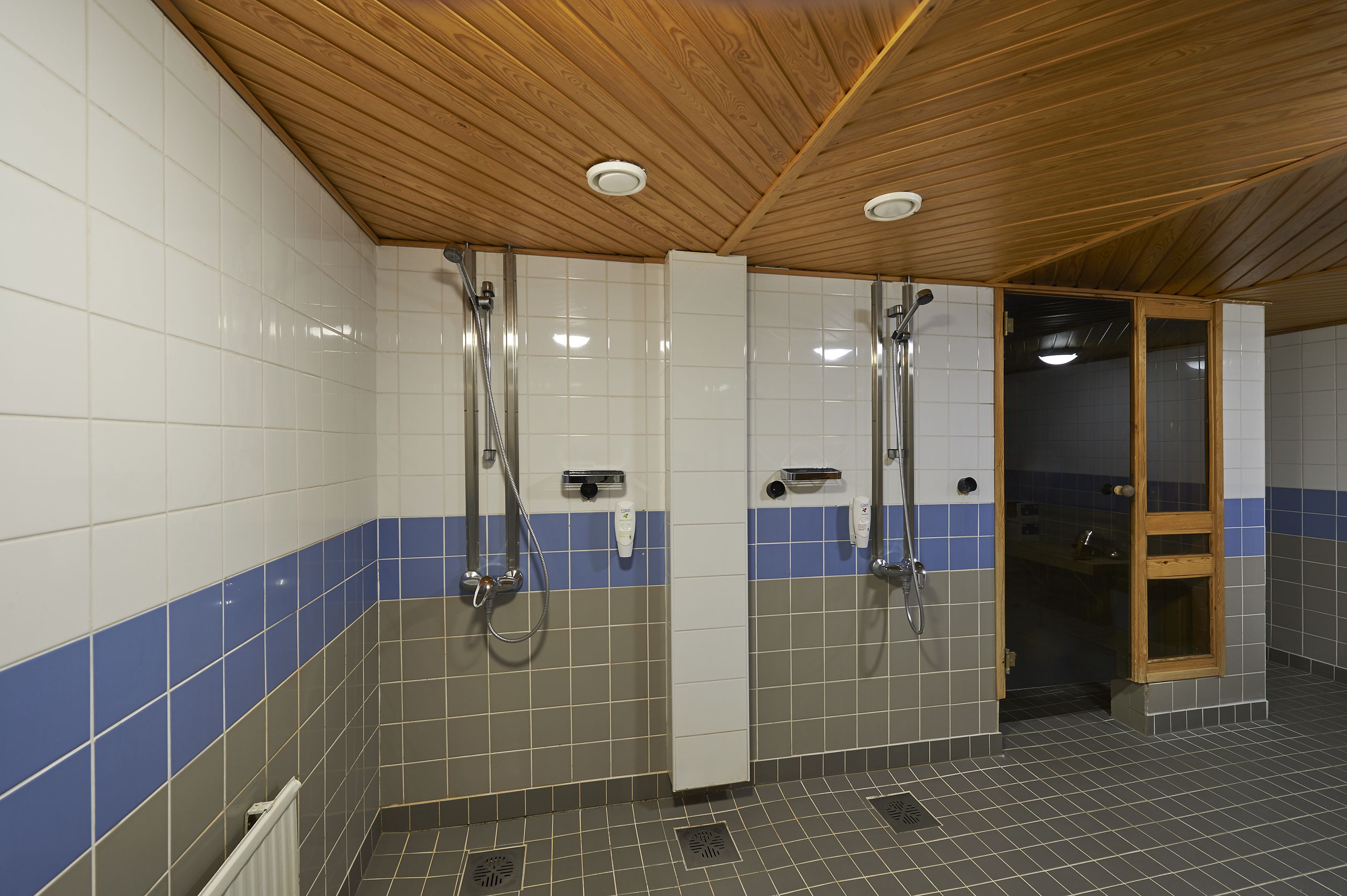 A tiled bathroom with wooden ceiling, two showerheads, and a glass door leading to another room.