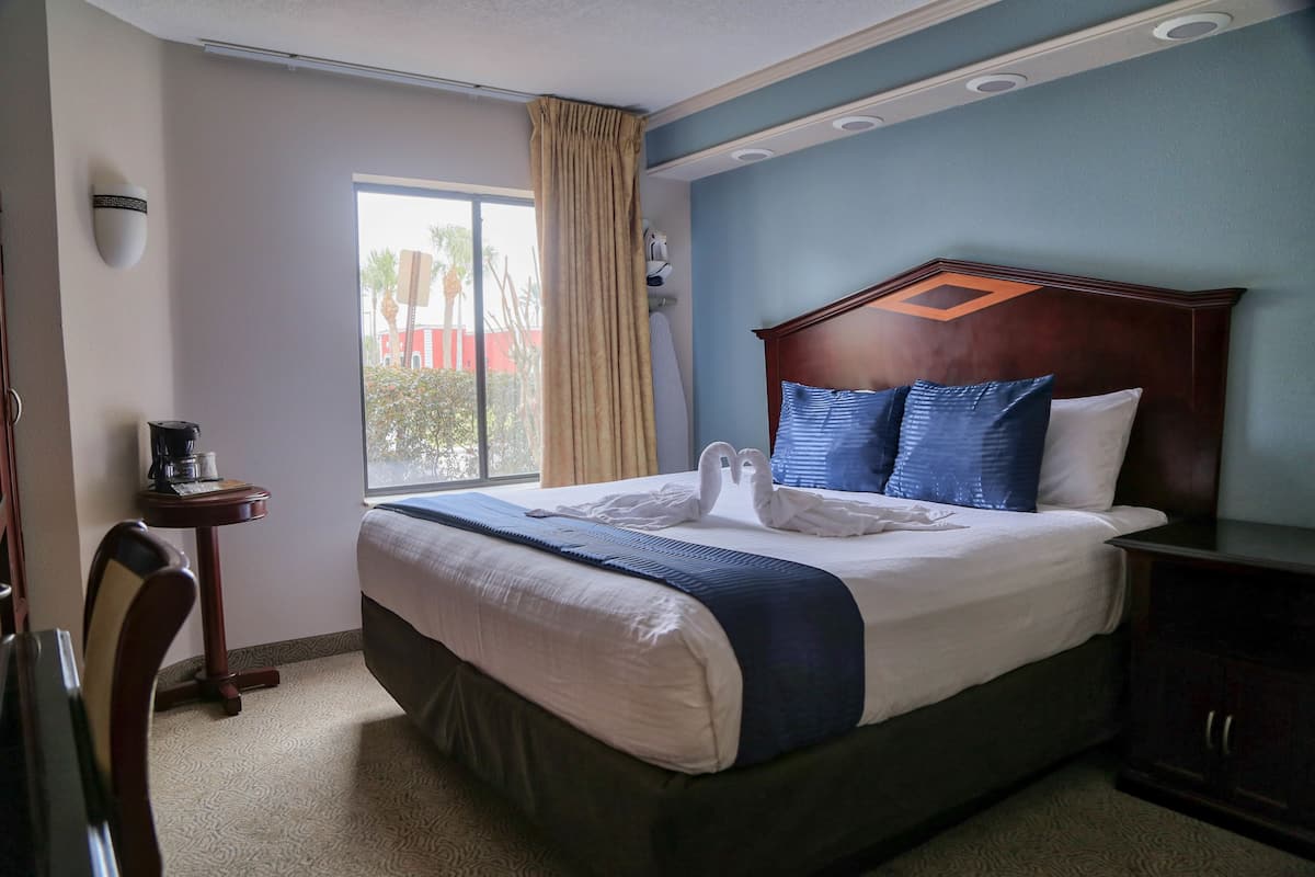 Habitación de hotel con una cama grande, cabecera de madera, ventana con cortinas, una mesita con cafetera y una mesita de noche.