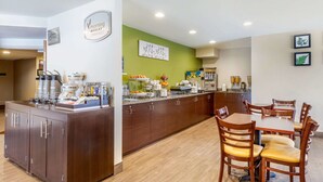 Ein Frühstücksbuffet mit Kaffeeautomaten, einer Obstauslage und einem Essbereich mit Holztischen und -stühlen.