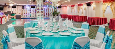 Un salón de banquetes con mesas redondas dispuestas para un evento formal, sillas con cintas azules y una zona de escenario con una pantalla y altavoces.