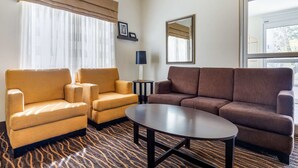 Ein Hotelzimmer mit zwei Sesseln, einem Sofa, einem Couchtisch, einer Lampe, einem Spiegel und einem fenster mit Vorhängen.