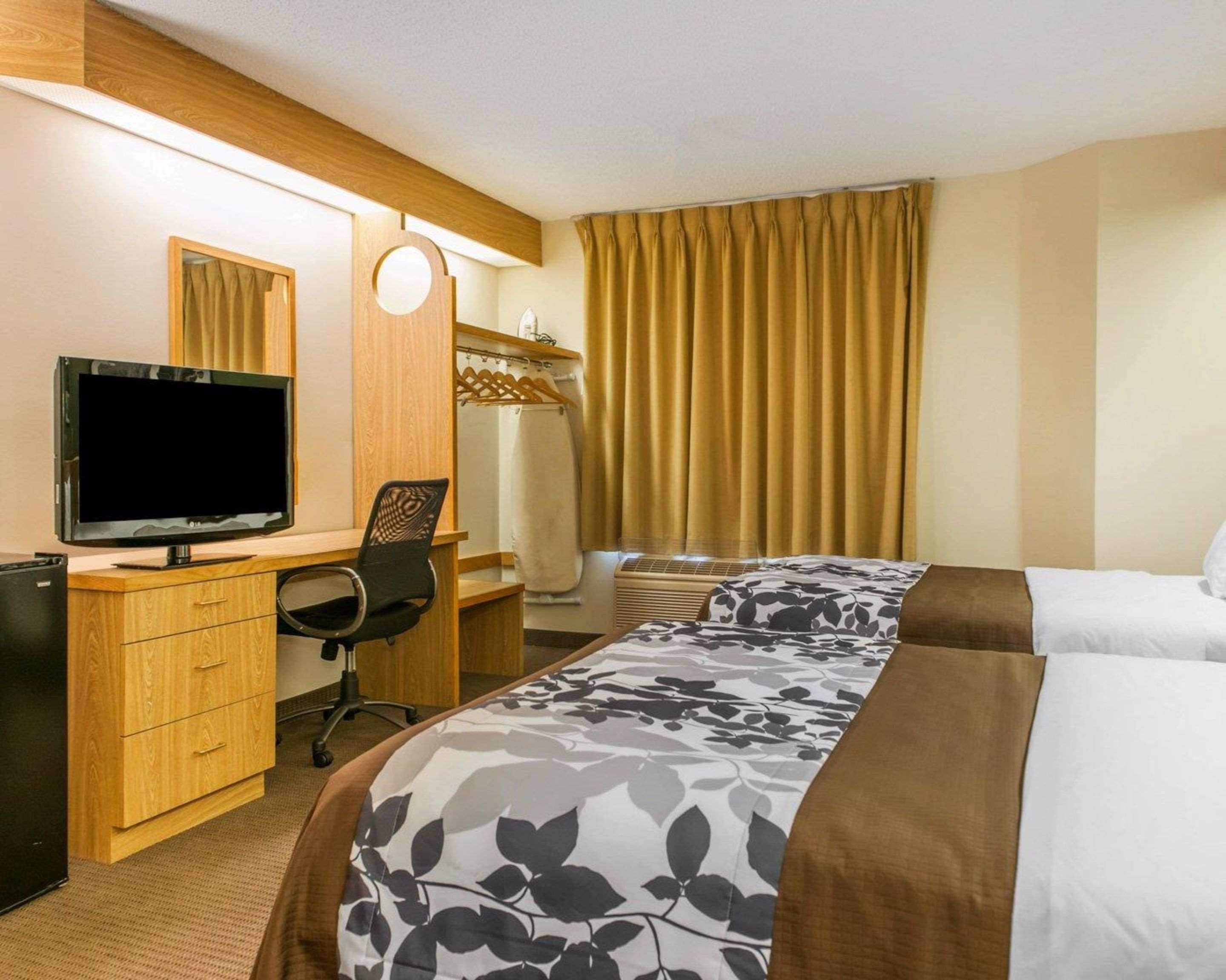 A hotel room with a TV, desk, chair, two beds, and a window with curtains.