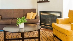 Una sala de estar con un sofá marrón, un sillón amarillo, una mesa redonda de centro con una planta y una chimenea con cuadros enmarcados arriba.