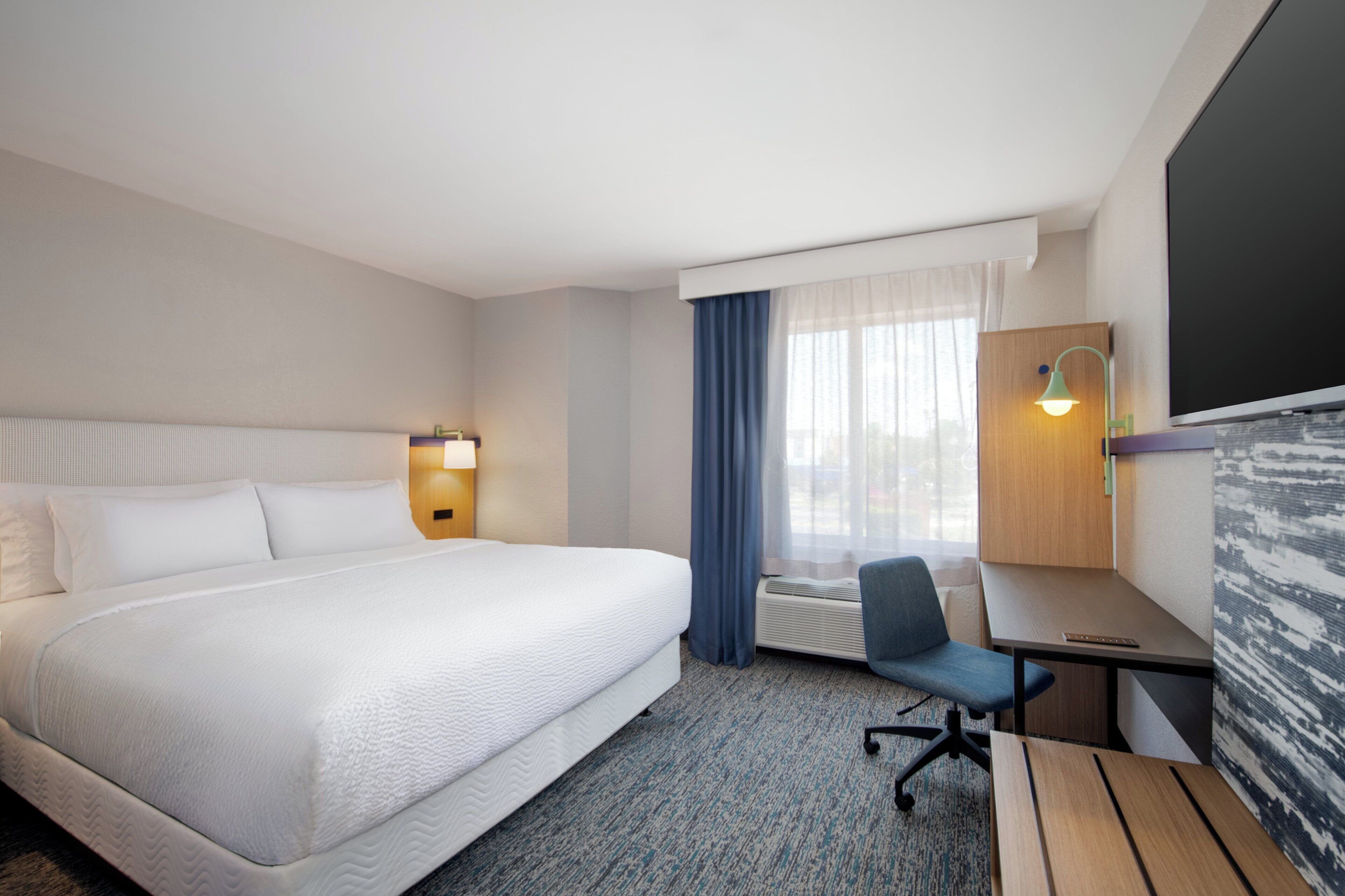 A hotel room with a large bed, a desk, a chair, a desk lamp, and a window with curtains.