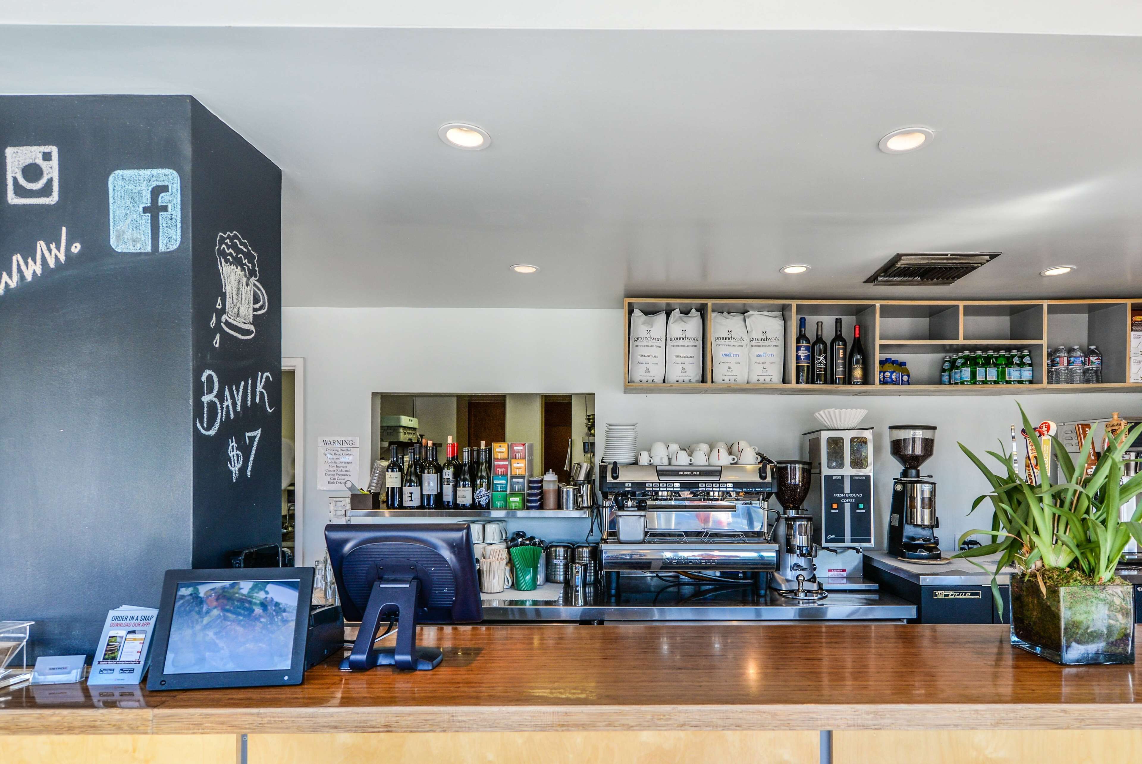 A coffee shop interior with a counter, espresso machines, shelves with bottled beverages, and a chalkboard with social media icons and a price.